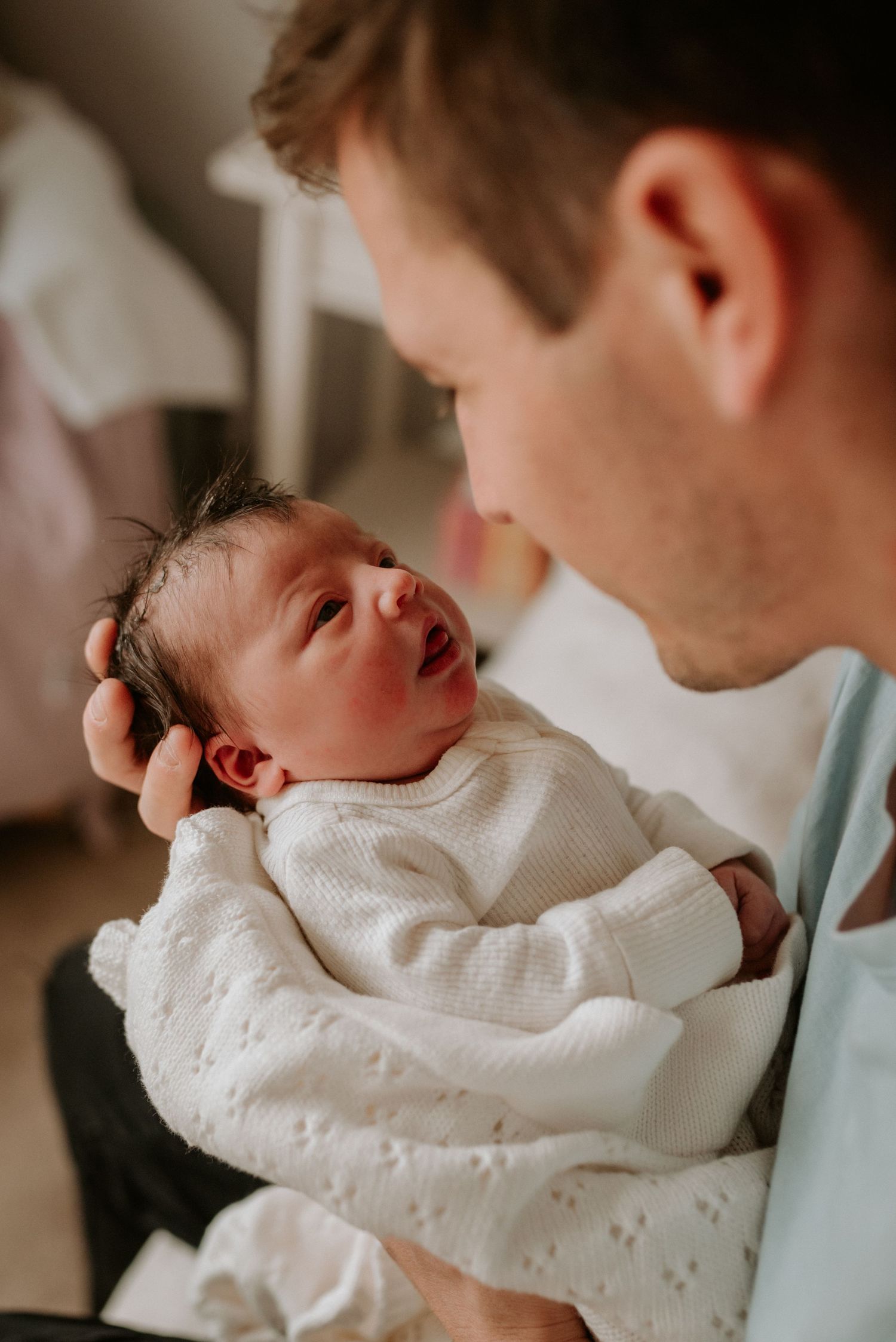 A baby in white clothing is cradled while being gazed at in soft natural light.