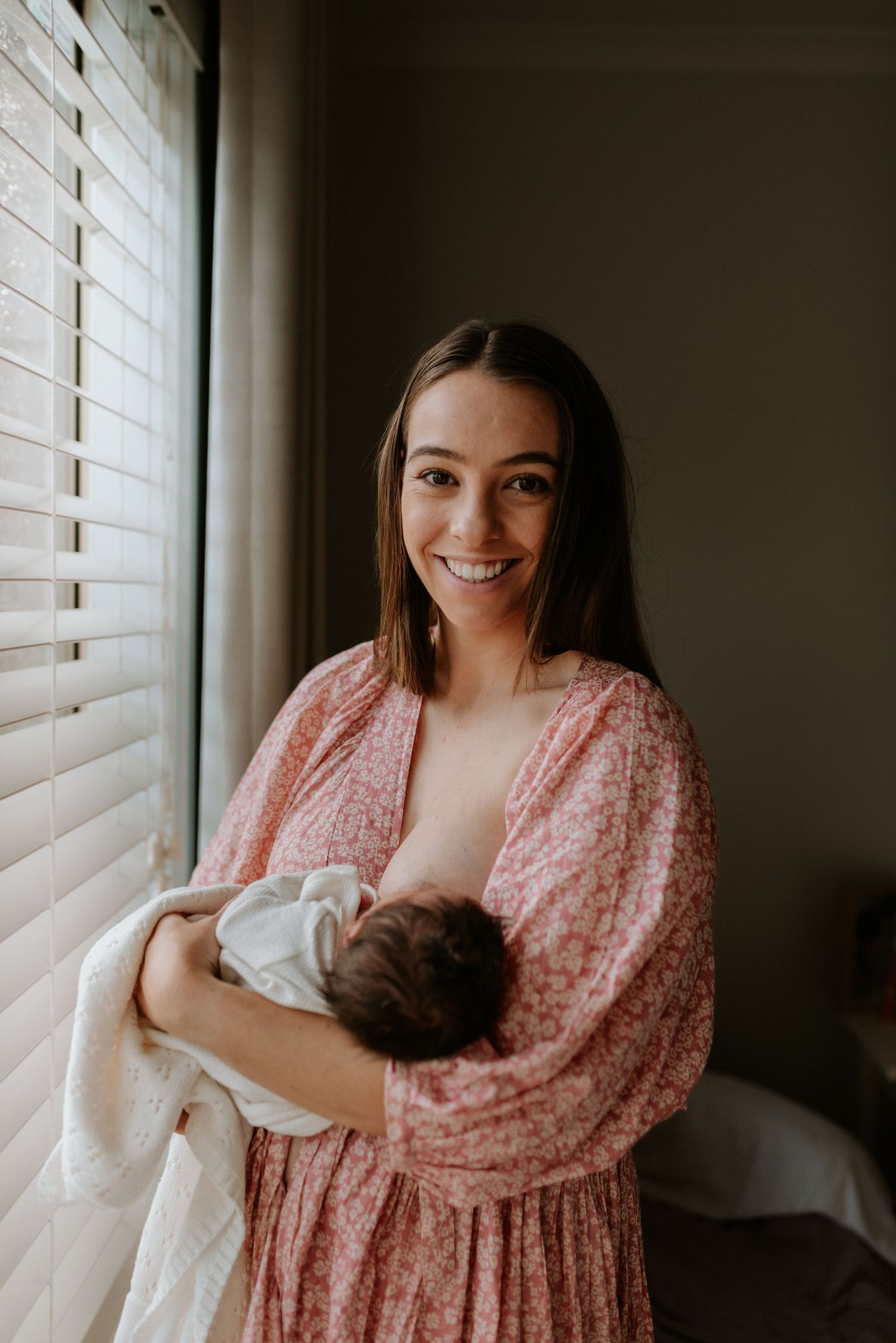 Mother in pink dress holding and nursing baby near window with venetian blinds.