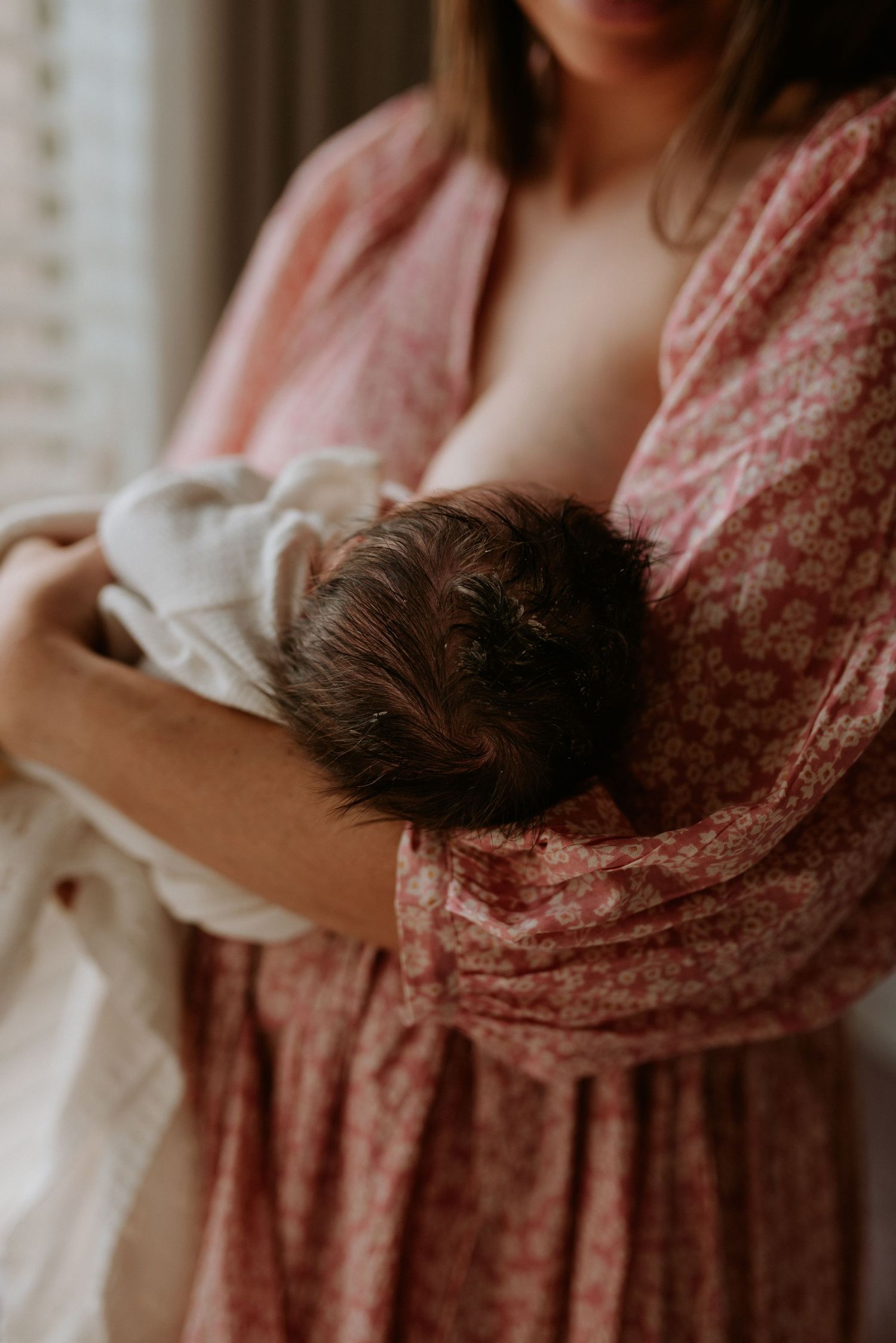 Tender moment captured in soft lighting showing person cradling baby against floral dress in muted pink tones.