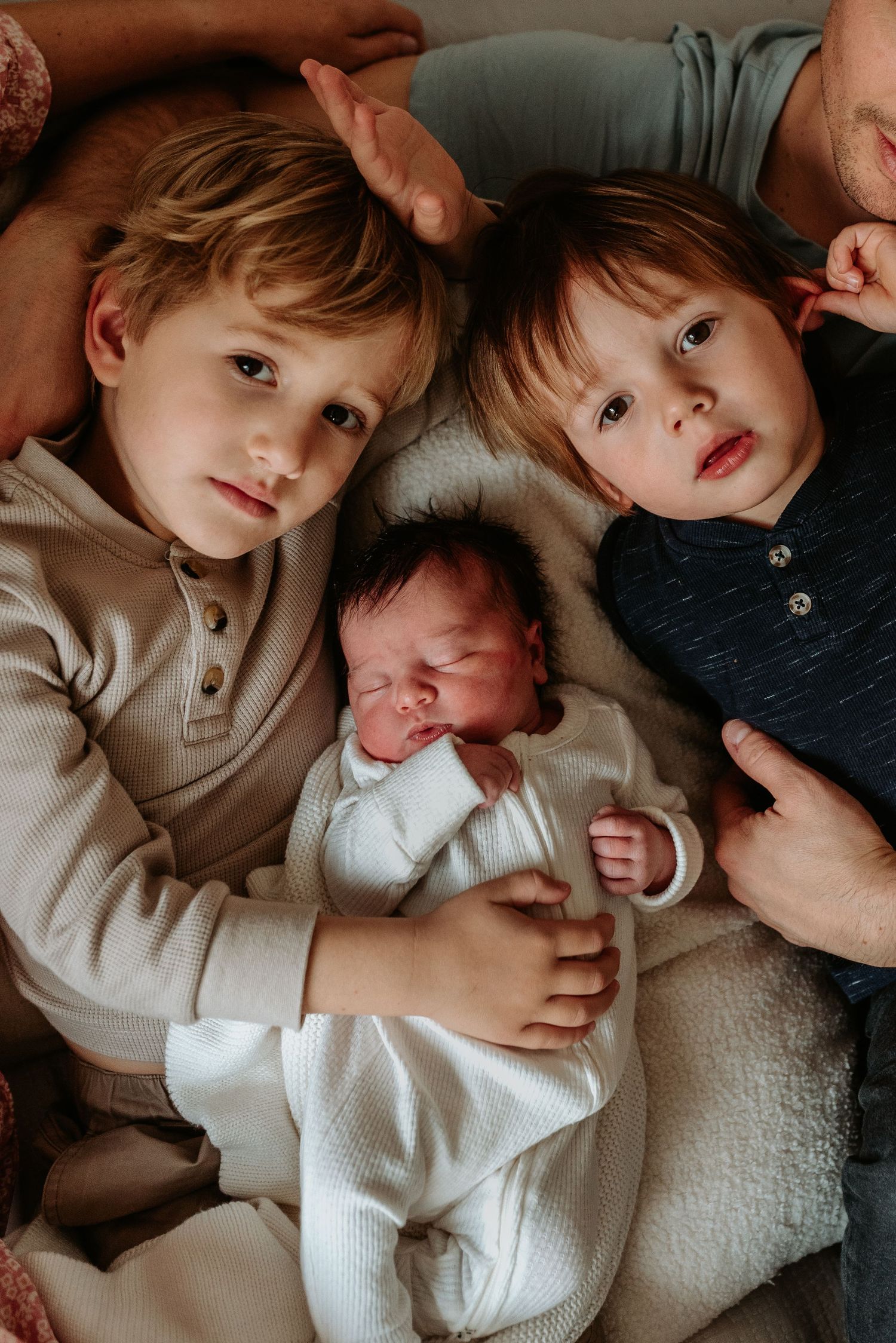 Two children lay together with a newborn baby wearing white on a light colored surface.