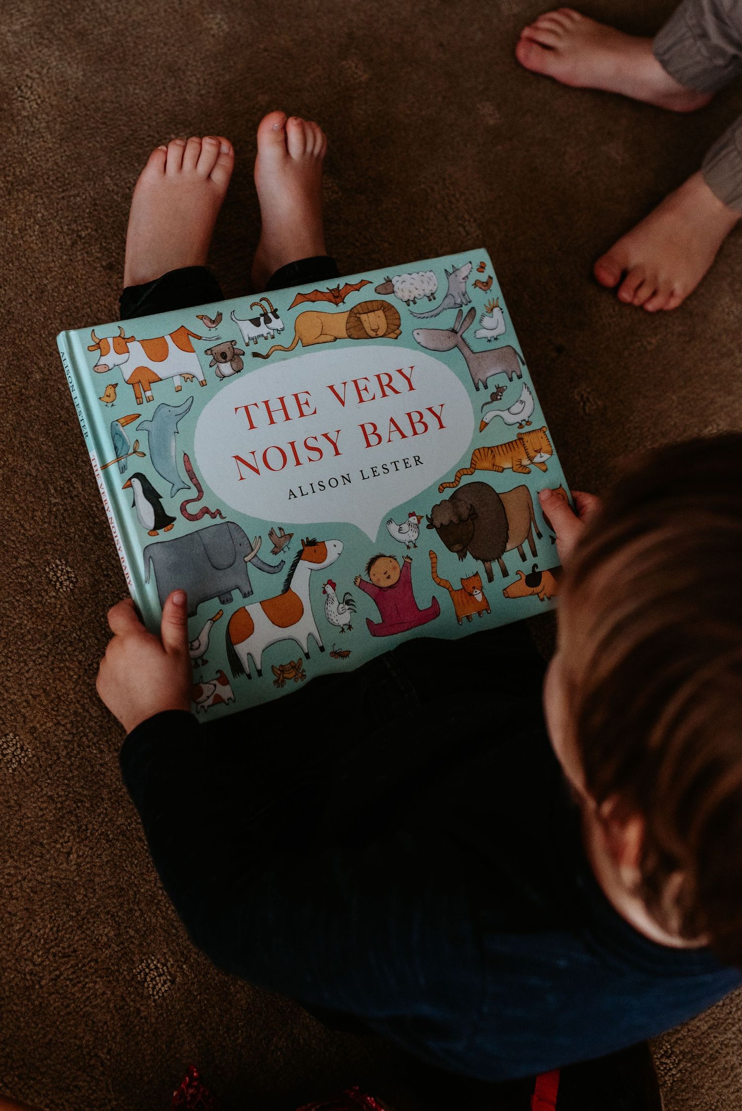 A child sits on a carpeted floor reading 'The Very Noisy Baby' children's book.