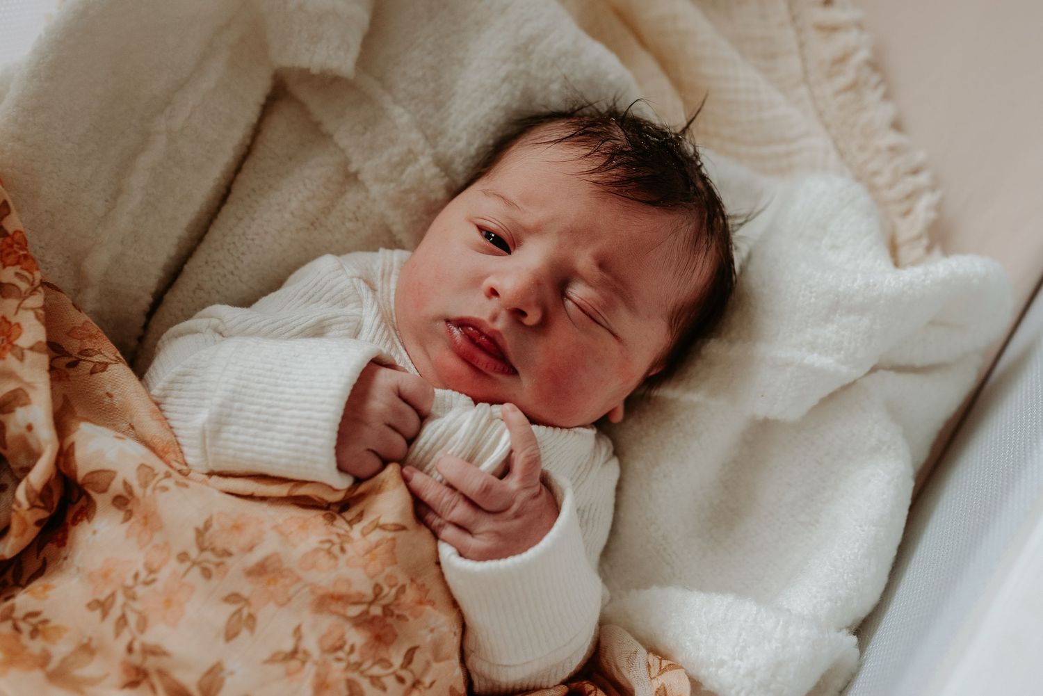A newborn baby in white clothing rests on a textured surface in warm lighting.