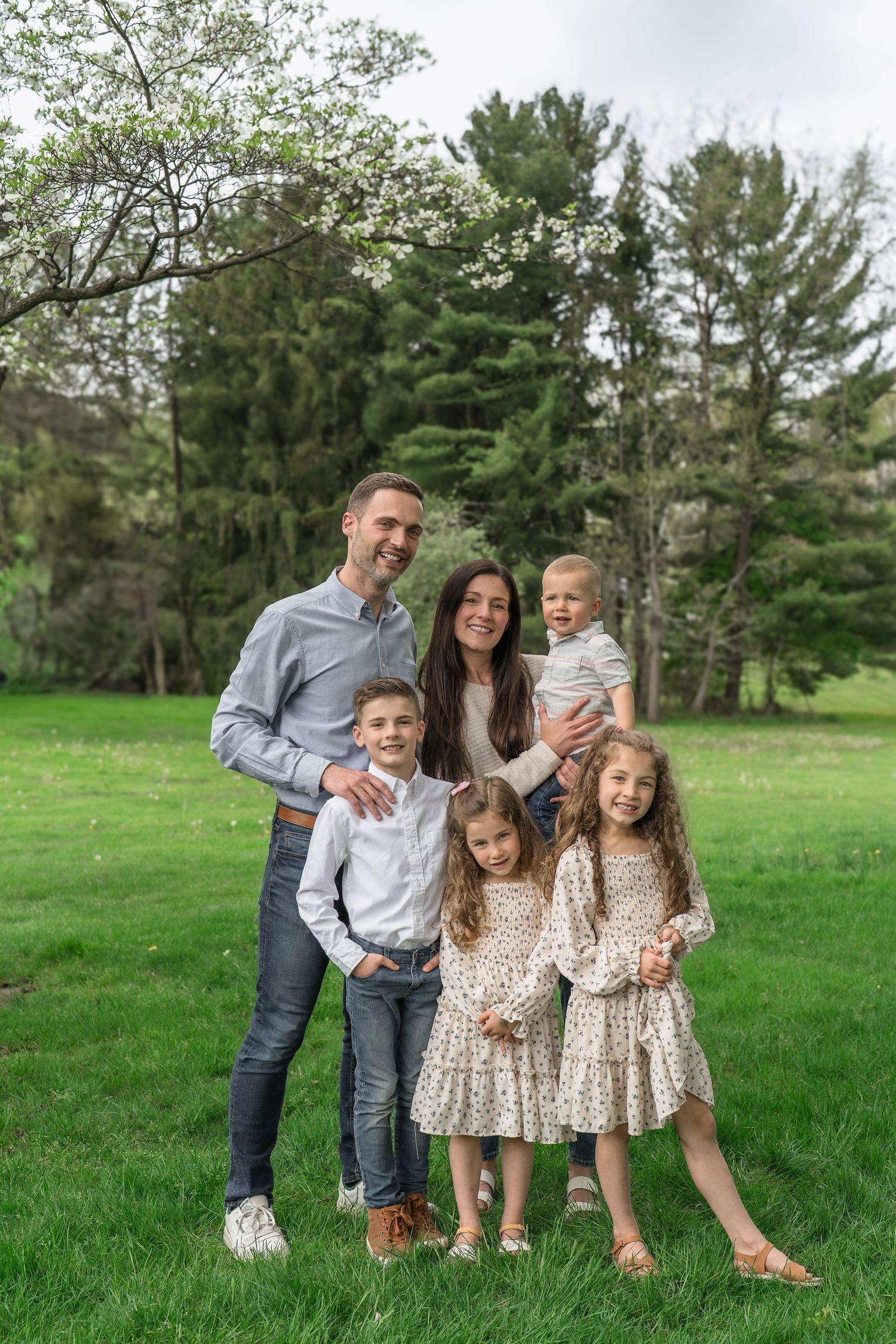 A group poses together outdoors on green grass with tall pine trees in the background on a spring day for an extended family session in Stow
