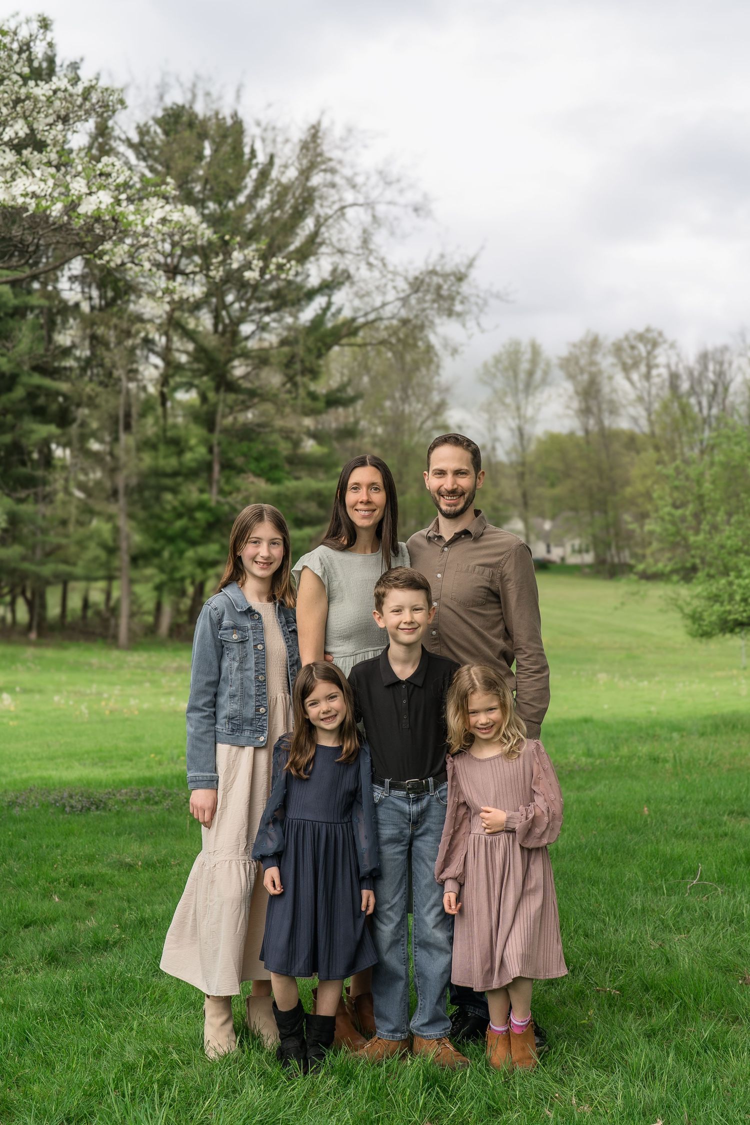 Family of six poses together outdoors in coordinated outfits on a green lawn with pine trees in the background at a Stow Ohio Family Session