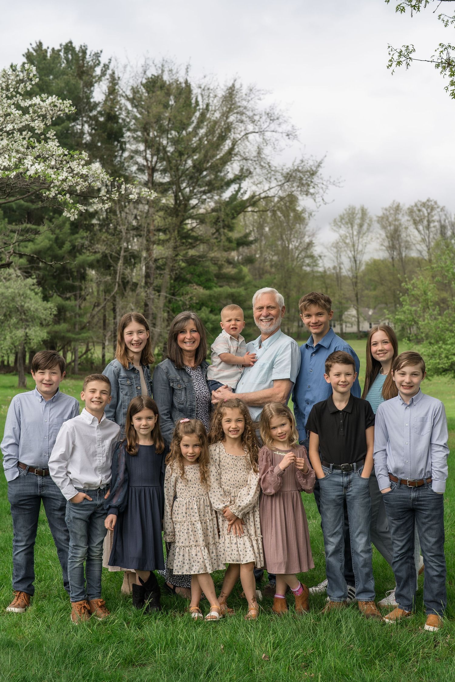 A multi-generational family group poses outdoors on green grass with trees in the background during springtime at a backyard in Stow, OH