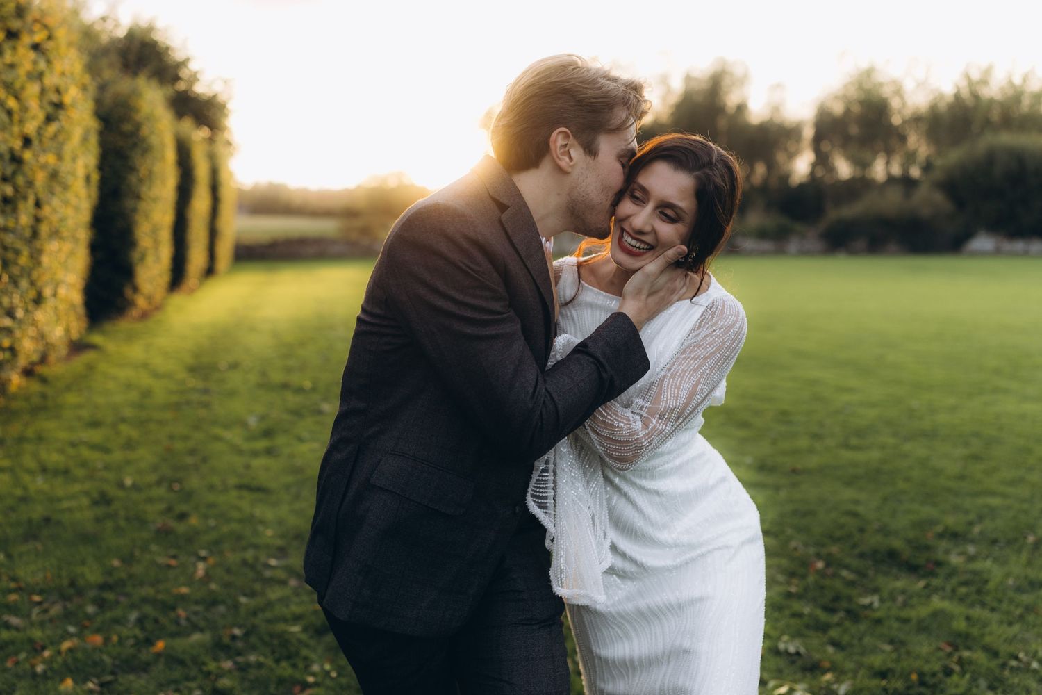 A romantic couple embraces on a grass lawn at sunset with manicured hedges in the background.