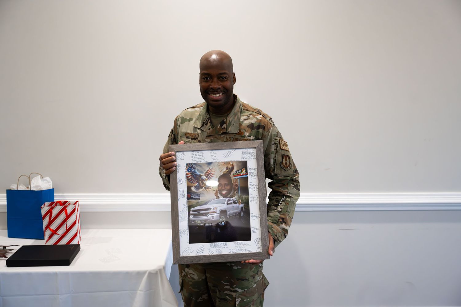 Service member in camouflage uniform displays framed photograph against white wall.