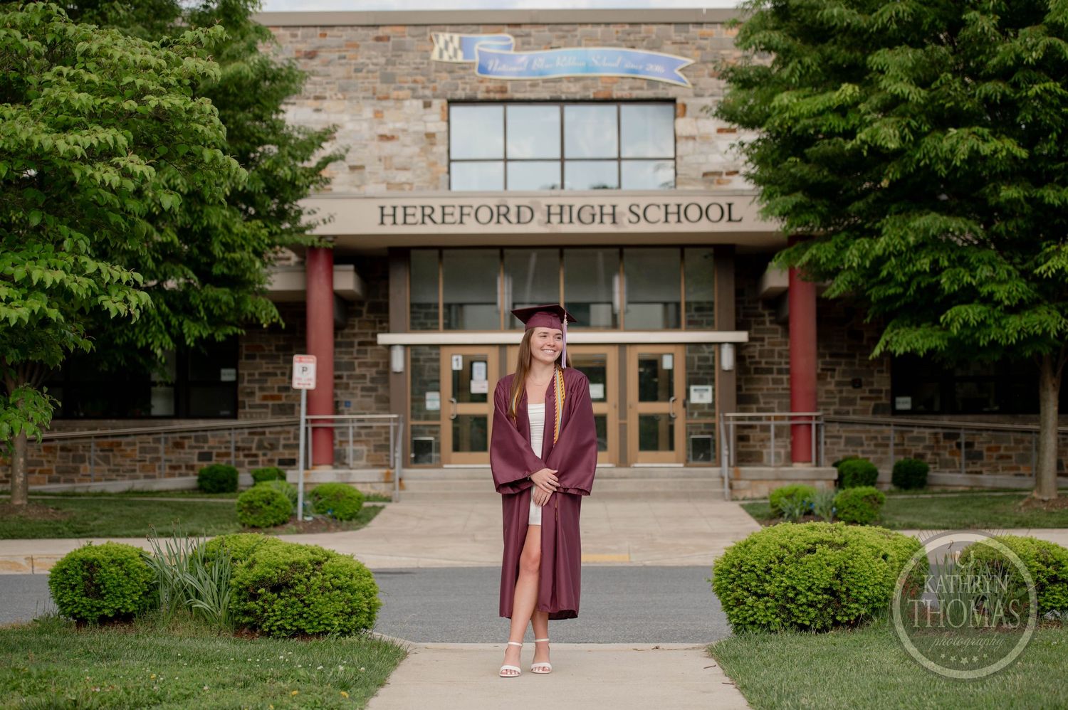 A graduate in burgundy cap and gown stands in front of Hereford High School's stone entrance on graduation day.