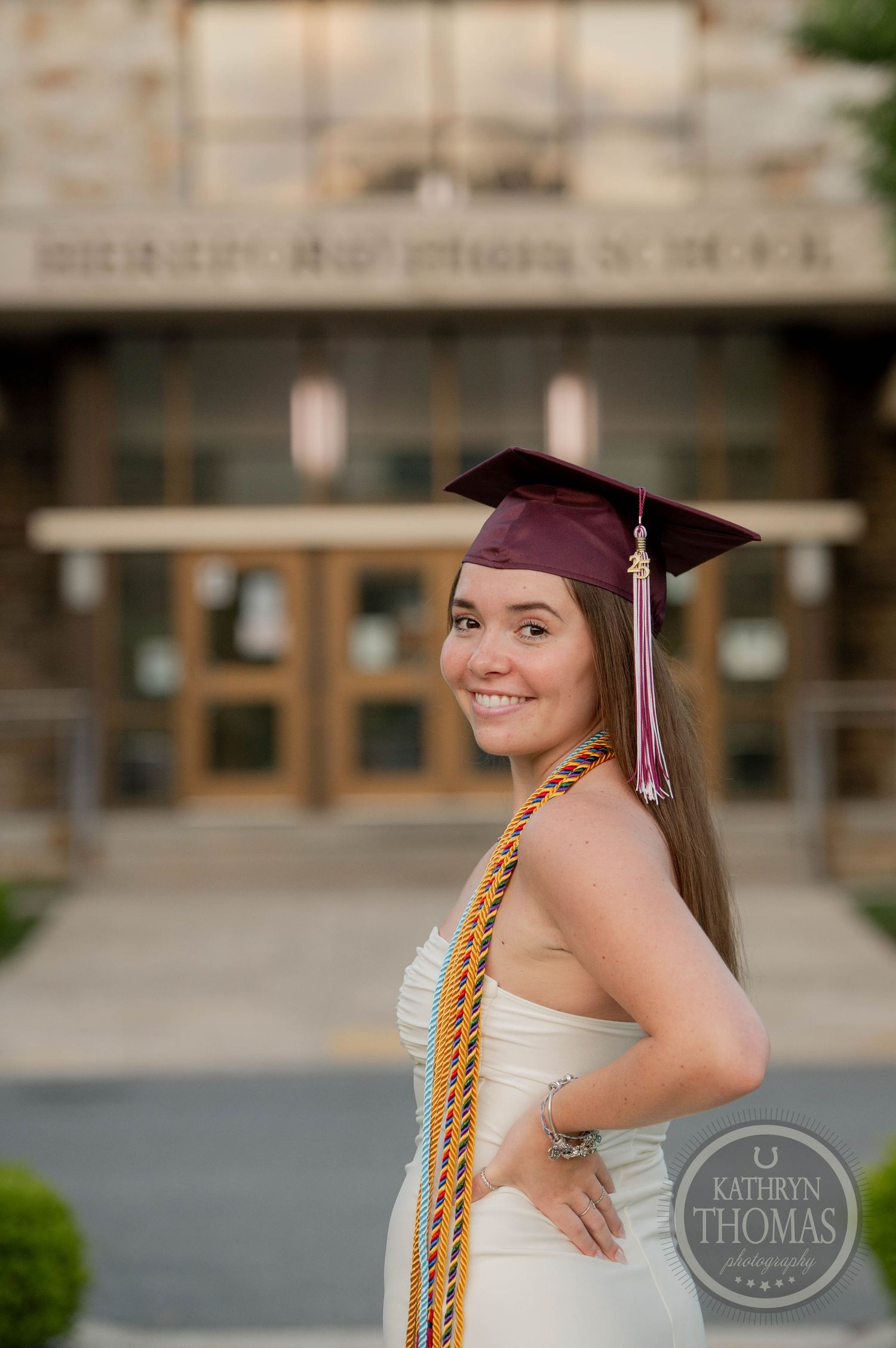Graduate in maroon cap and gold honor cords poses in front of campus building in white dress for graduation photos.