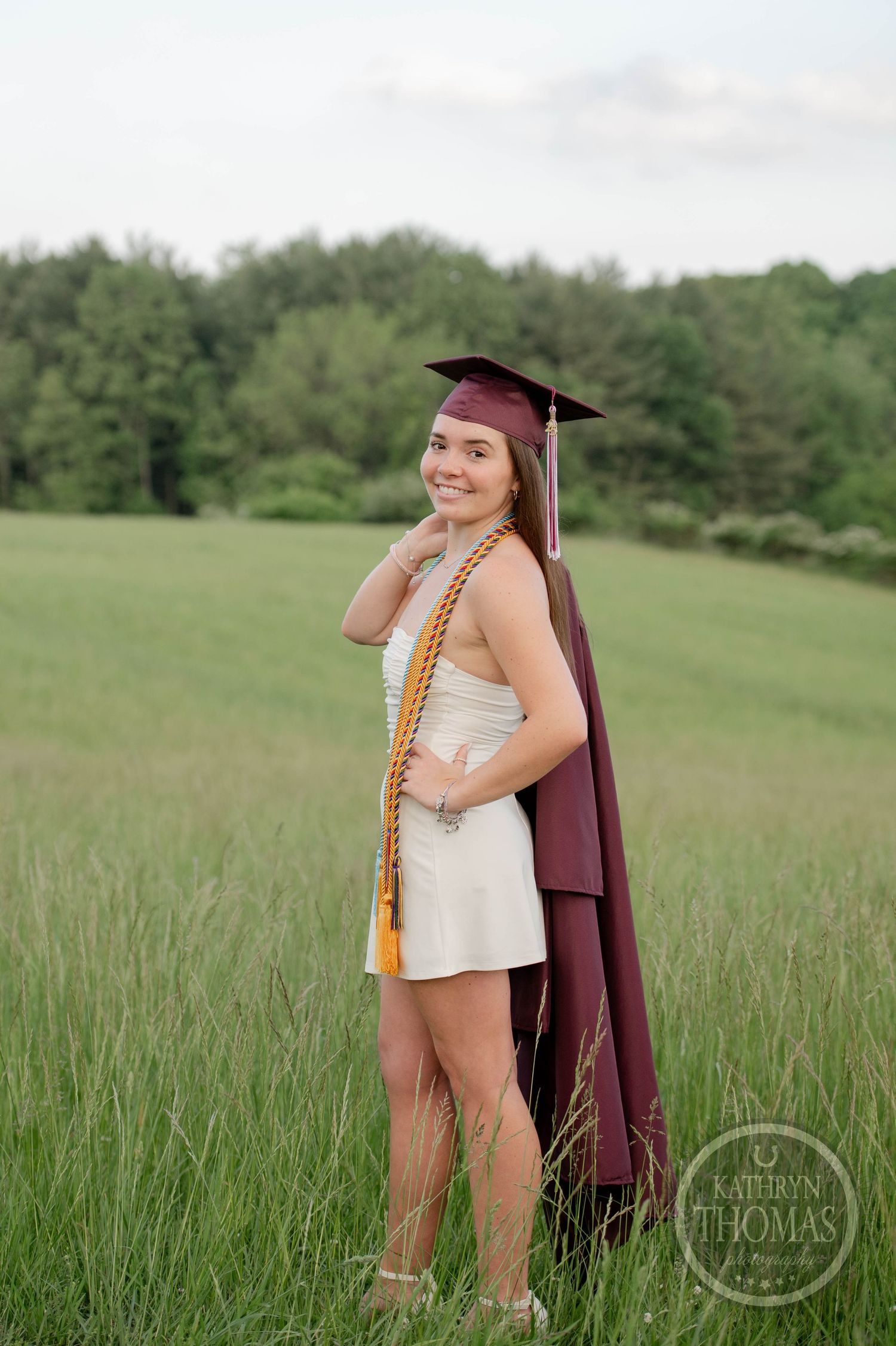 Graduate in maroon cap and gown stands in a sunny green field during outdoor graduation photo session.