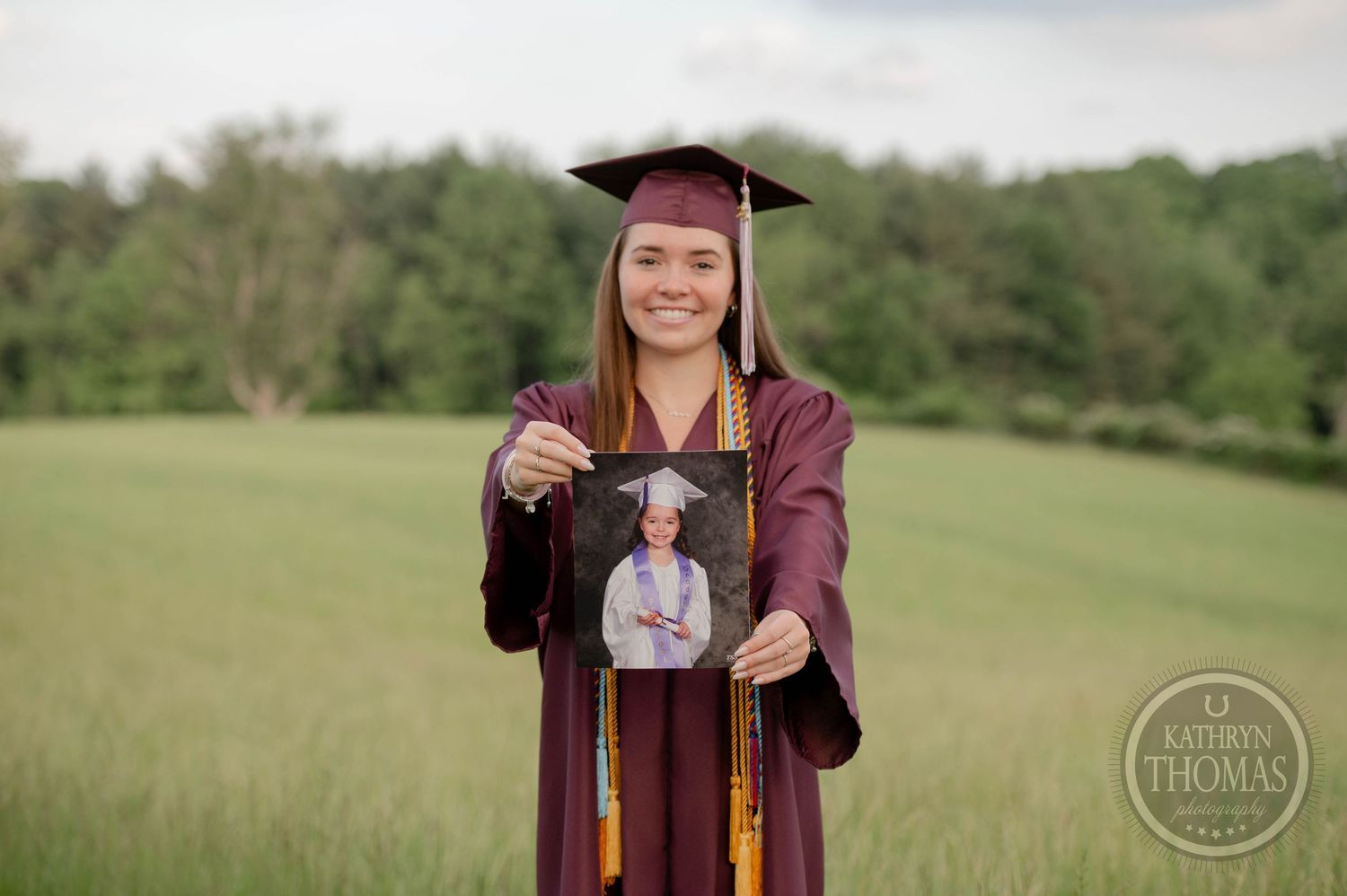 Graduate in burgundy cap and gown holds up a framed photo while standing in a green field.