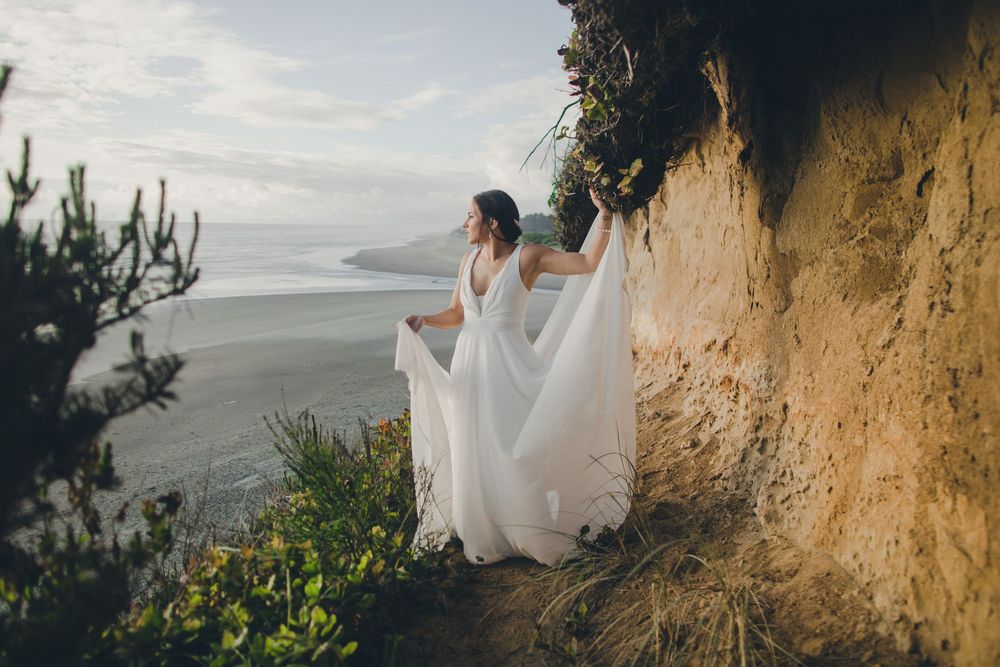 A figure in a flowing white dress poses dramatically against a steep coastal cliff at sunset.