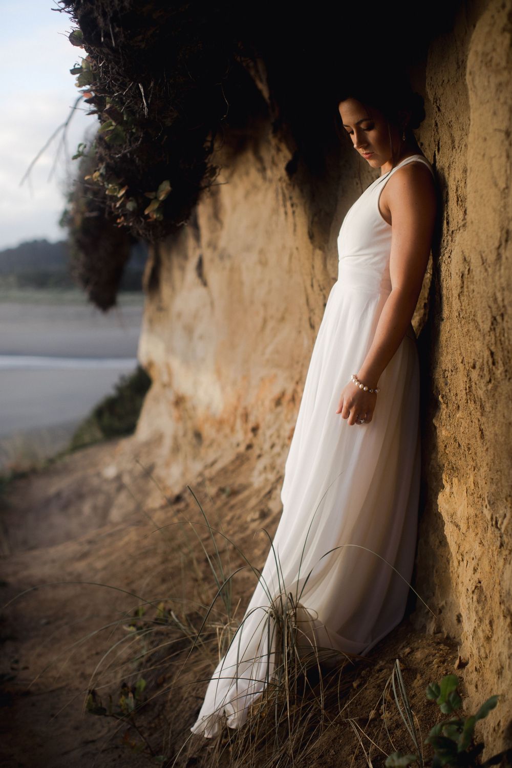 Person in flowing white dress leans against weathered cliff wall at sunset along coastal path.