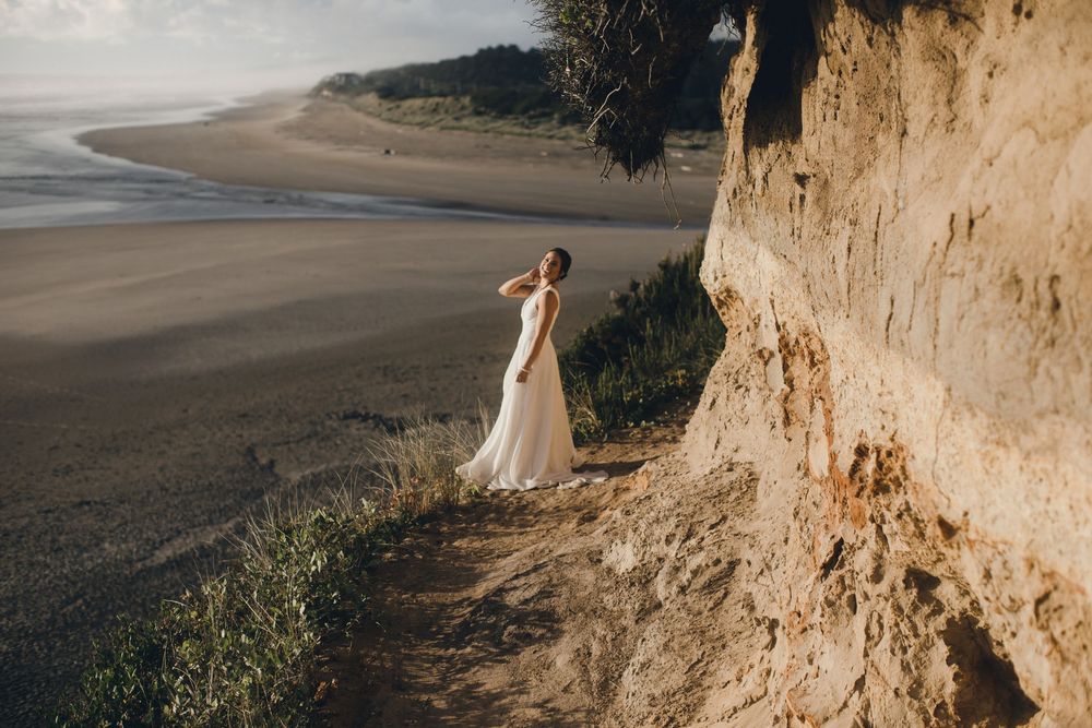 A wedding dress clad figure walks along a cliff edge path with sweeping ocean and beach views below.