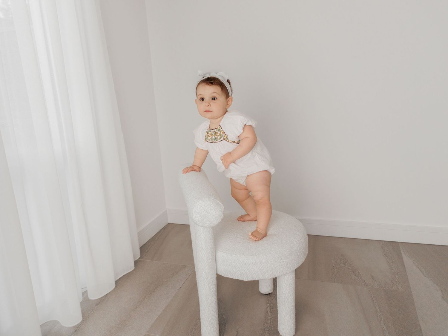 Baby in white outfit plays with wooden letters against light backdrop in minimalist studio setting.
