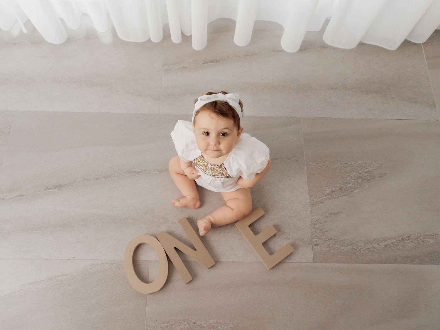 Baby in white outfit explores wooden letters on light hardwood floor with white curtains in background.