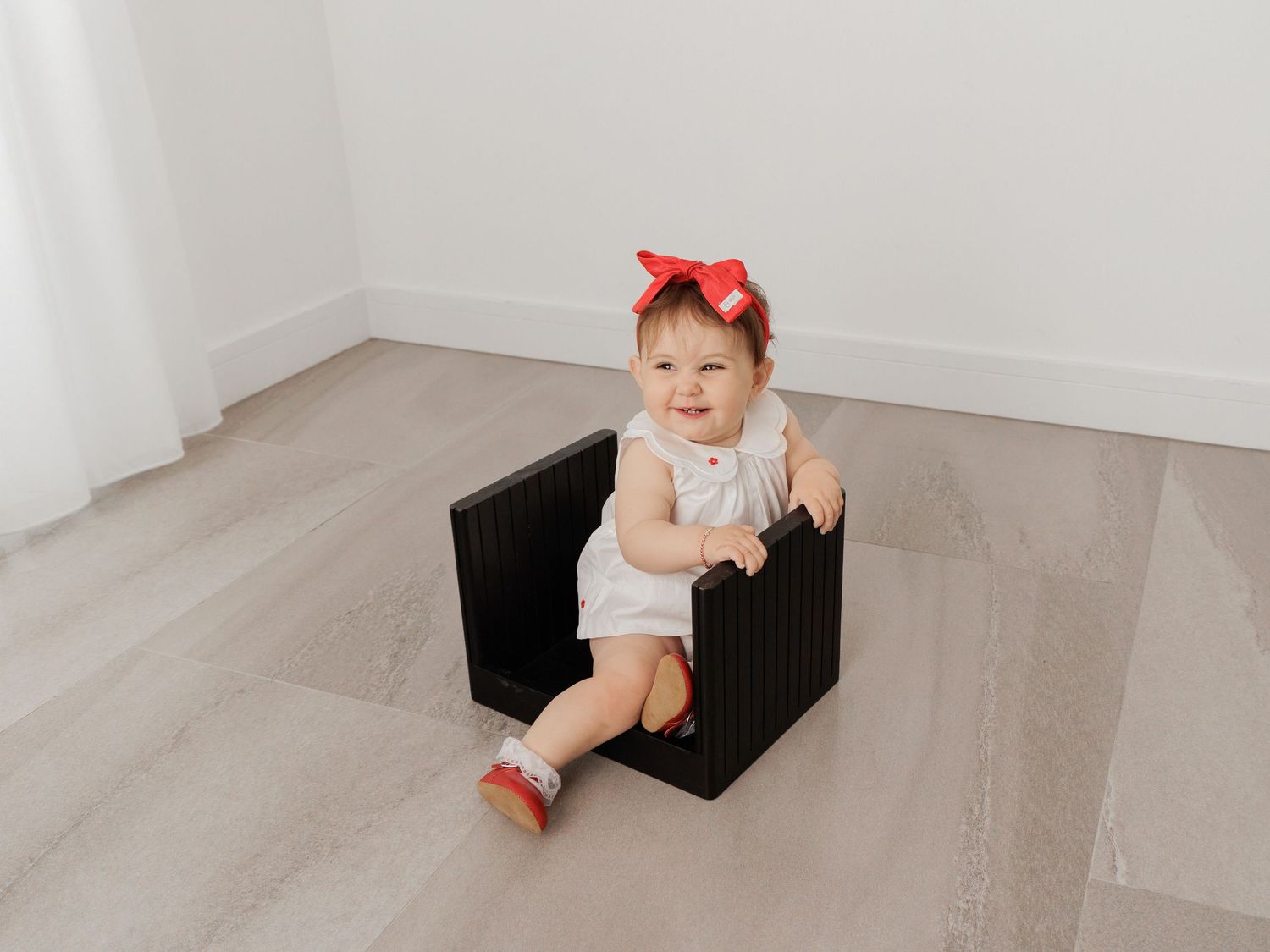 A baby in white clothing and red bow sits on a black woven cube ottoman on a gray wood floor.