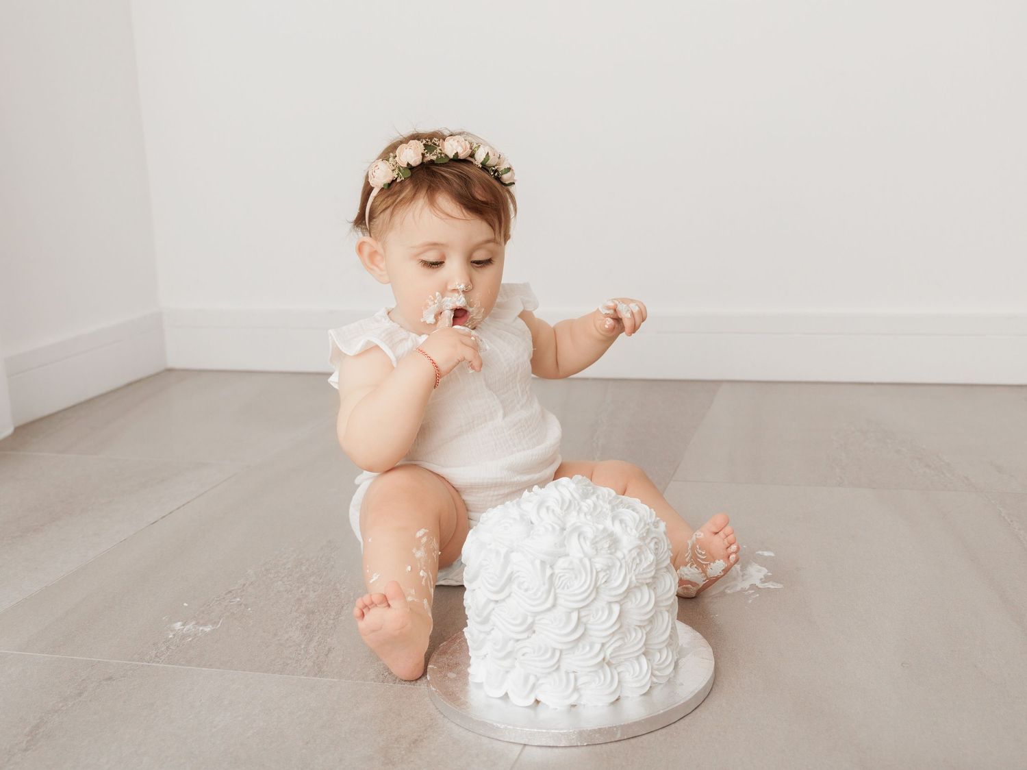 Baby wearing white outfit sitting on floor next to white birthday smash cake during photoshoot.