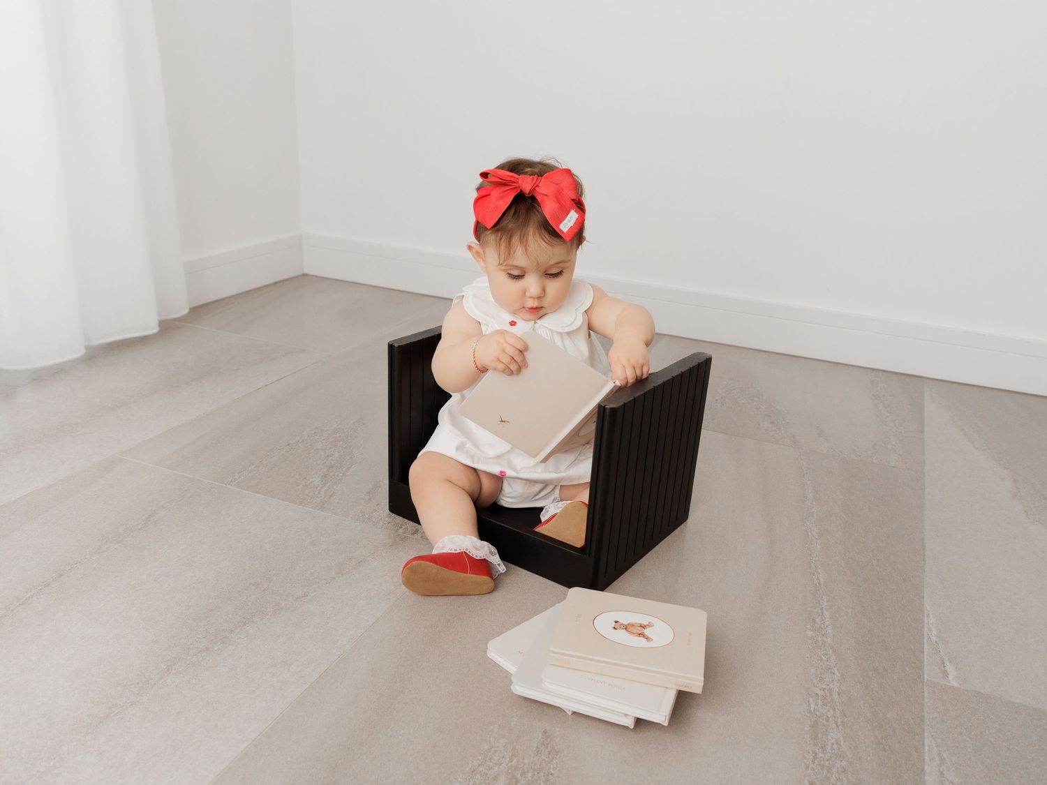 Baby in white onesie sits on wooden chair against white wall with book.
