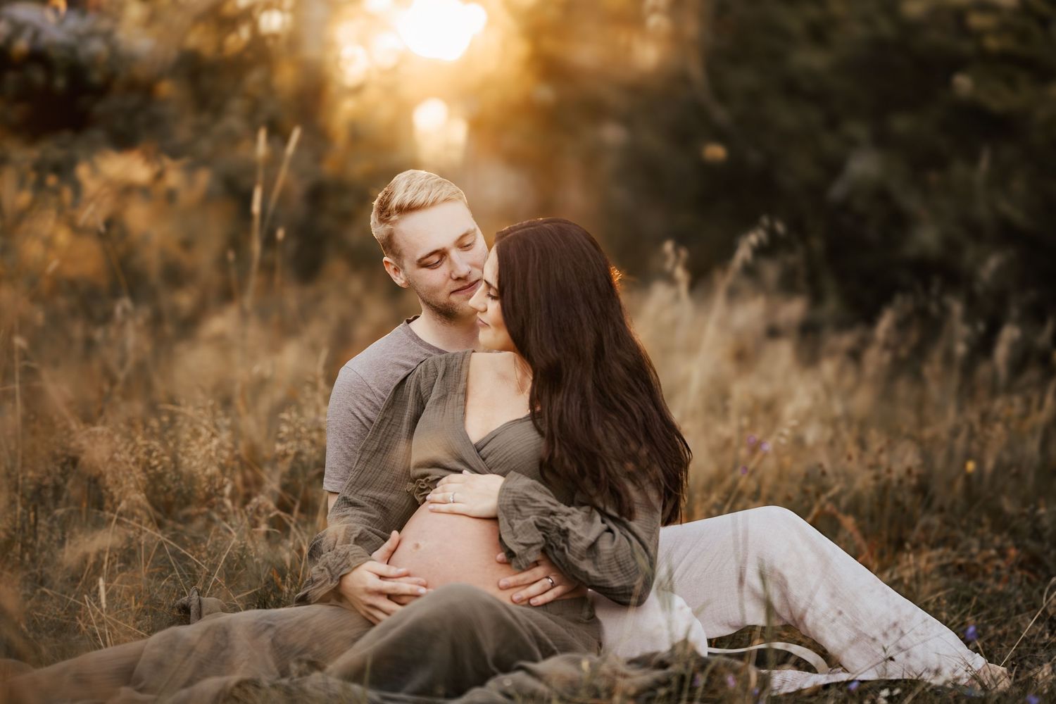 A woman in a flowy white maternity dress cradles her pregnant belly while standing among delicate spring blossoms.