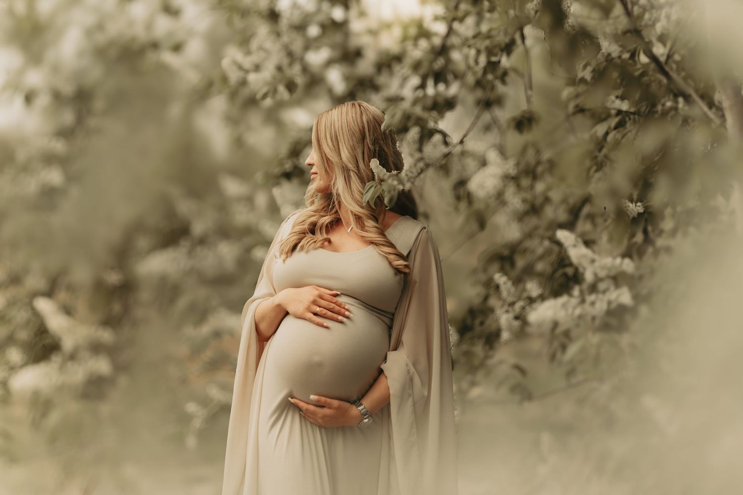 A woman in a flowy white maternity dress cradles her pregnant belly while standing among delicate spring blossoms.