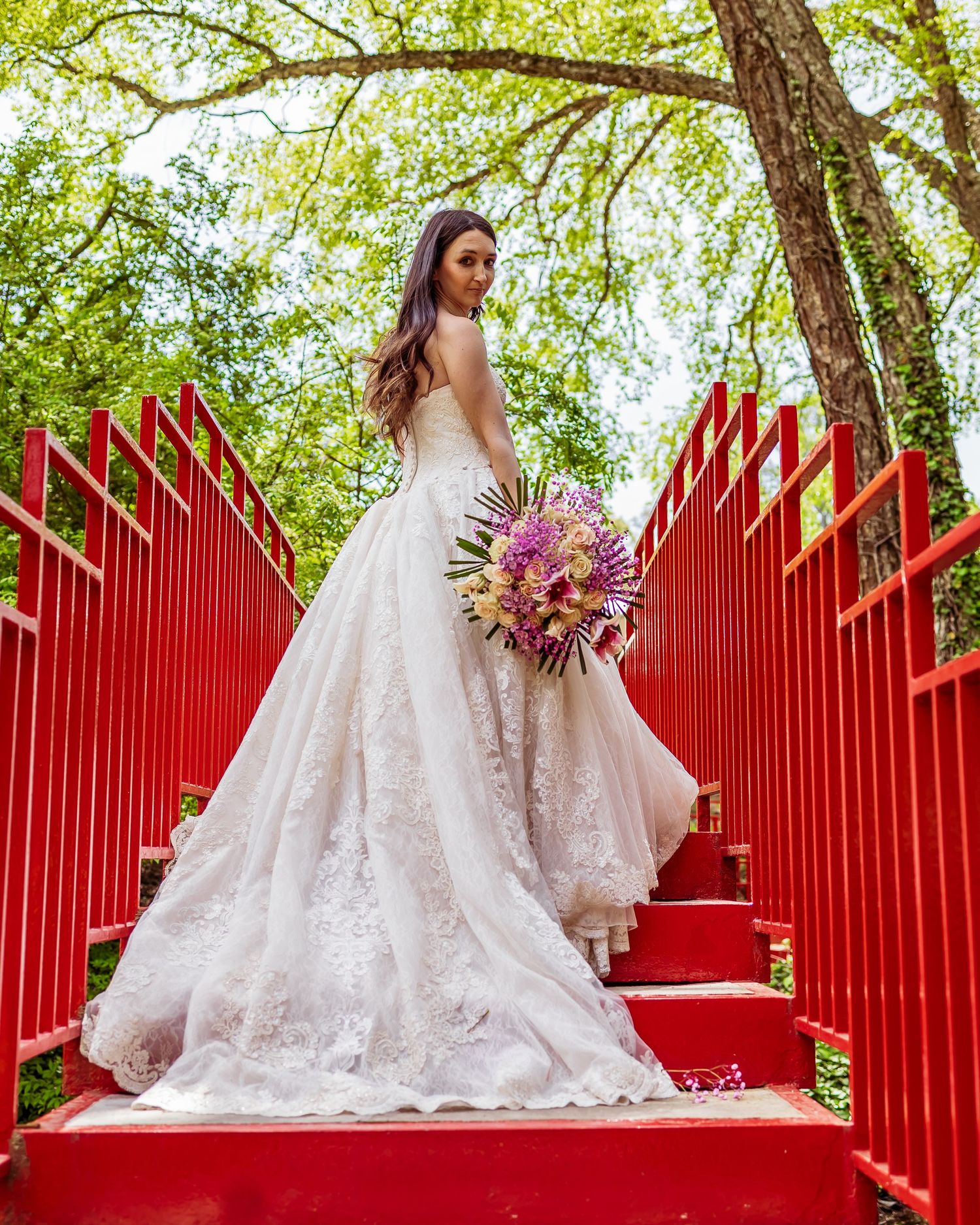 Bride in lace wedding gown standing on red stairs surrounded by greenery while holding pink bouquet on her special day.