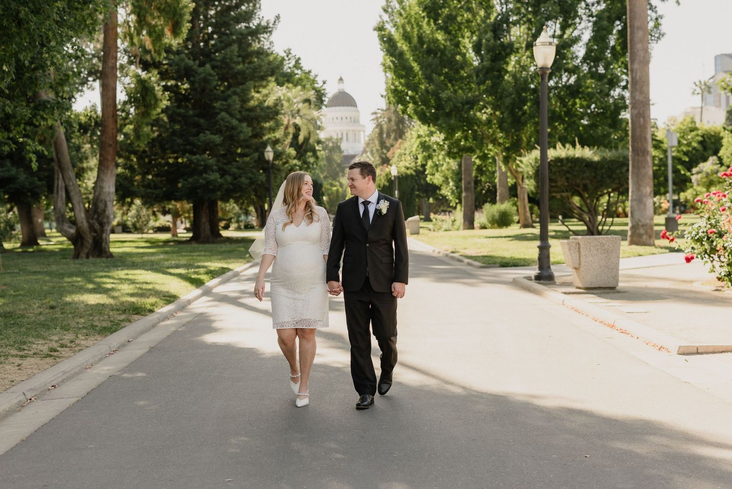A couple walks together on a tree-lined pathway in formal wedding attire on a sunny day.