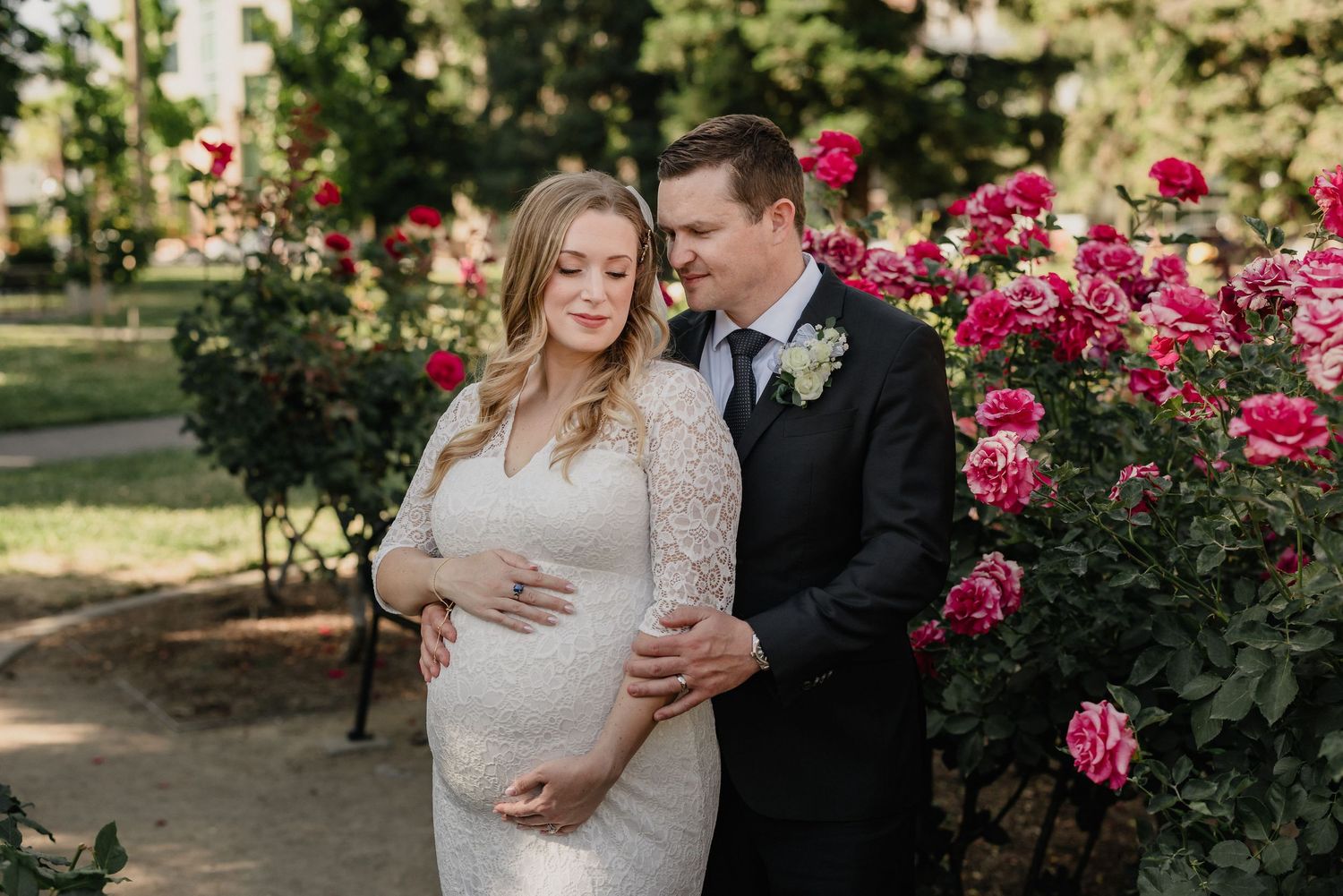 Expecting couple in formal attire embracing among blooming pink rose bushes in a garden.
