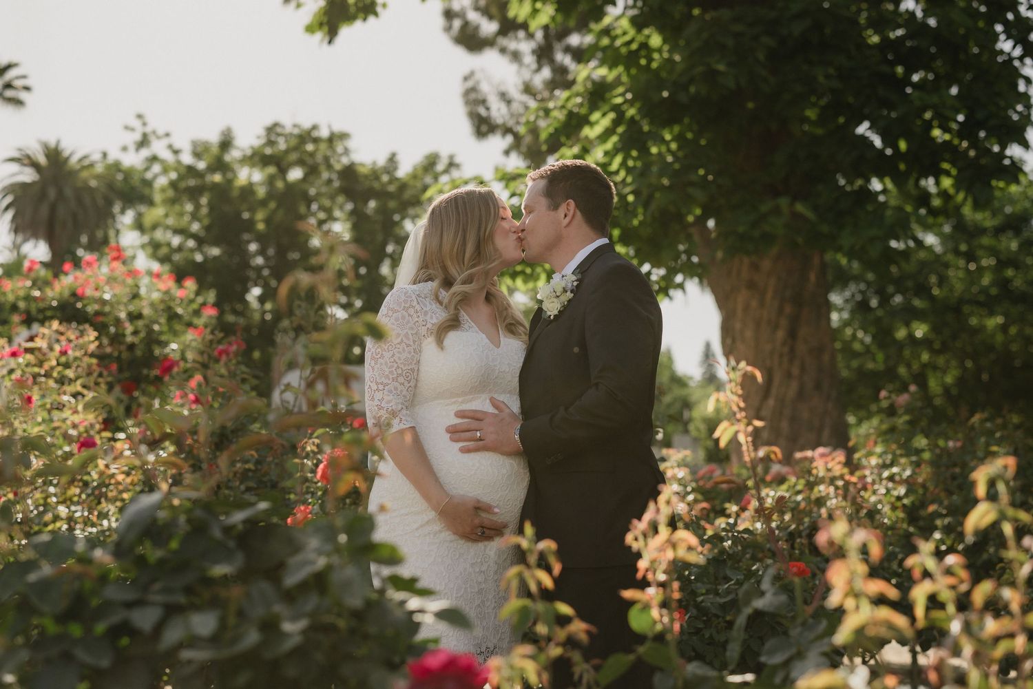 Newlyweds share a tender moment in a garden with roses and golden sunlight filtering through the trees.
