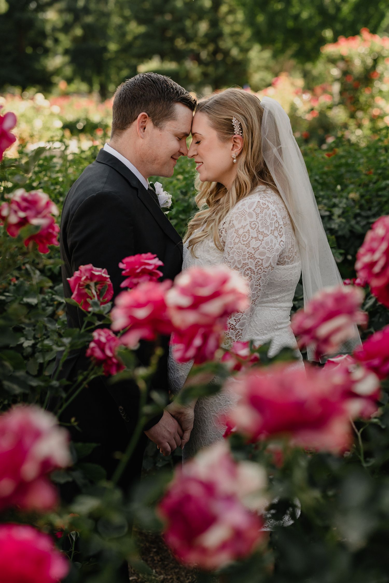 A romantic wedding photo of a couple sharing an intimate moment among blooming pink roses in a garden setting.