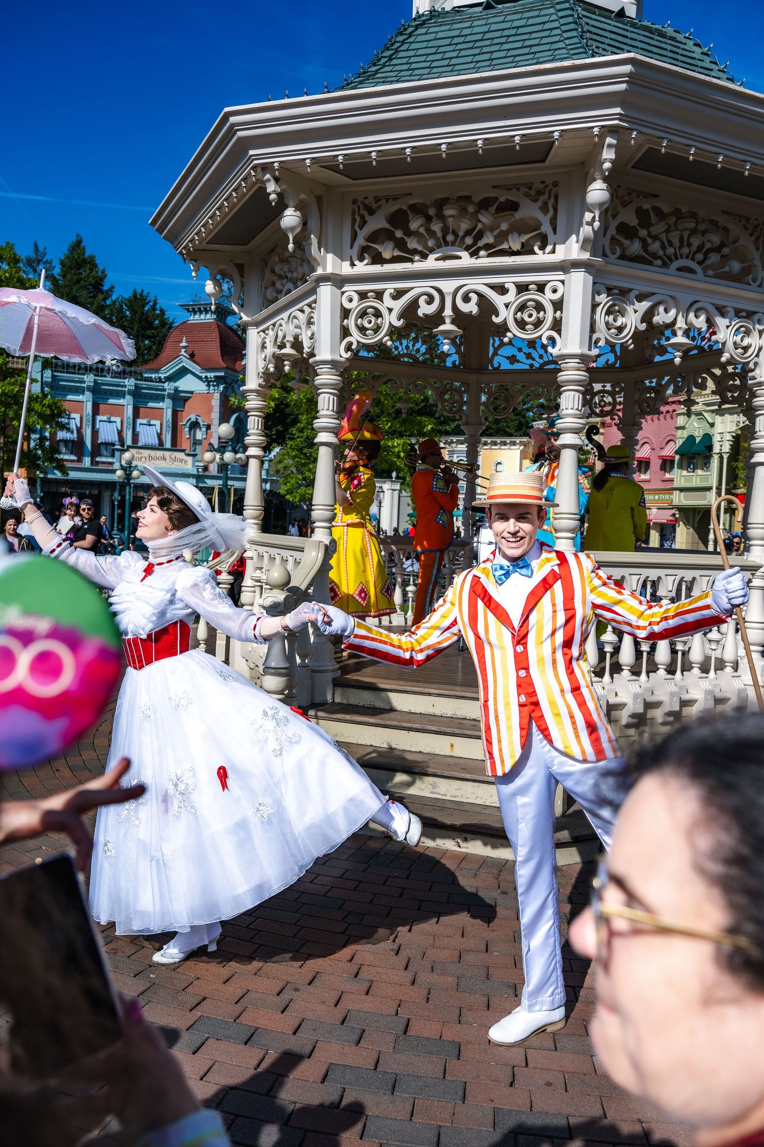 Vintage performers in Victorian-era costumes dance and entertain guests near an ornate gazebo at Disneyland theme park.