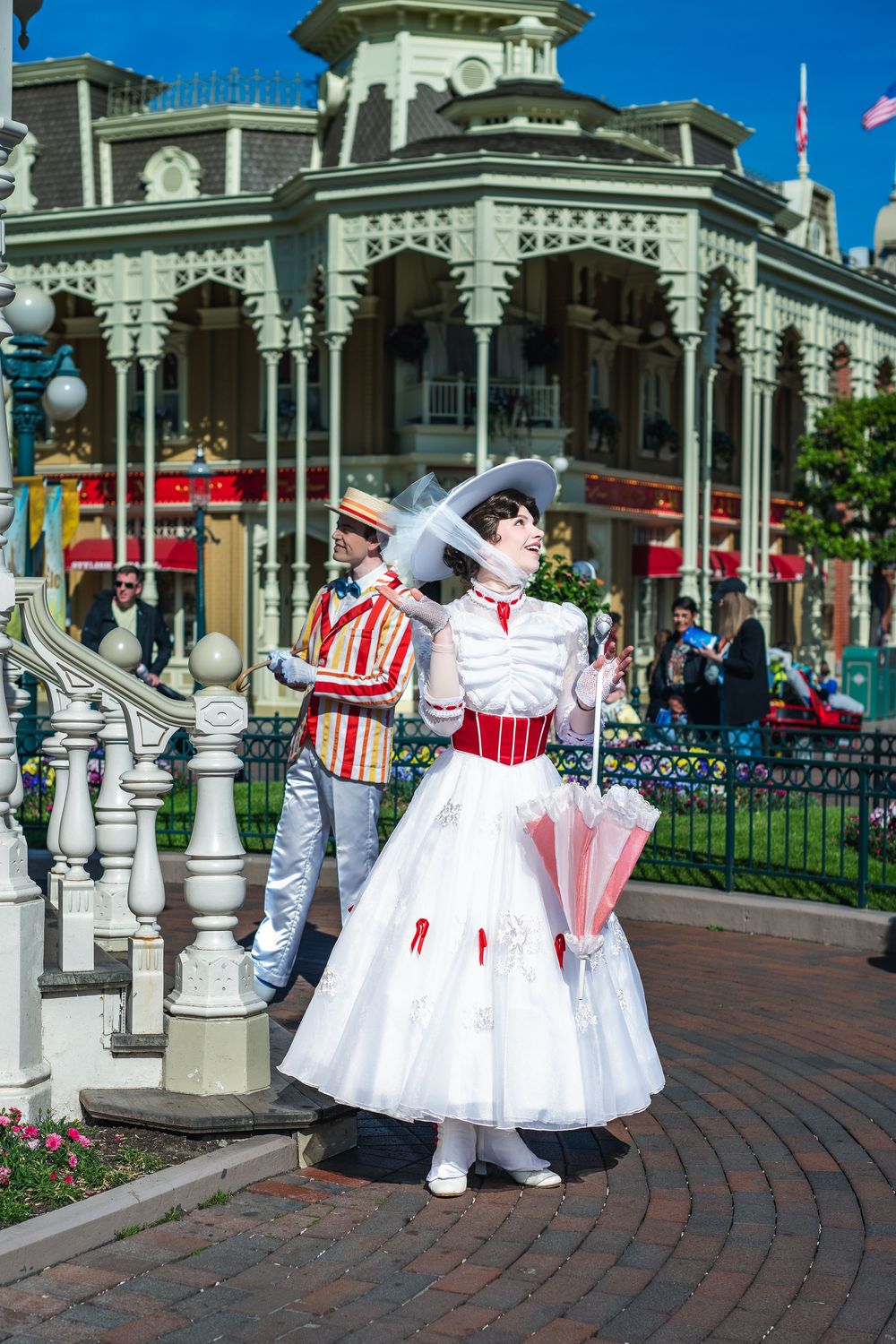 A performer in a Victorian-style white and red dress with parasol stands on a brick path at Disney's Main Street USA.