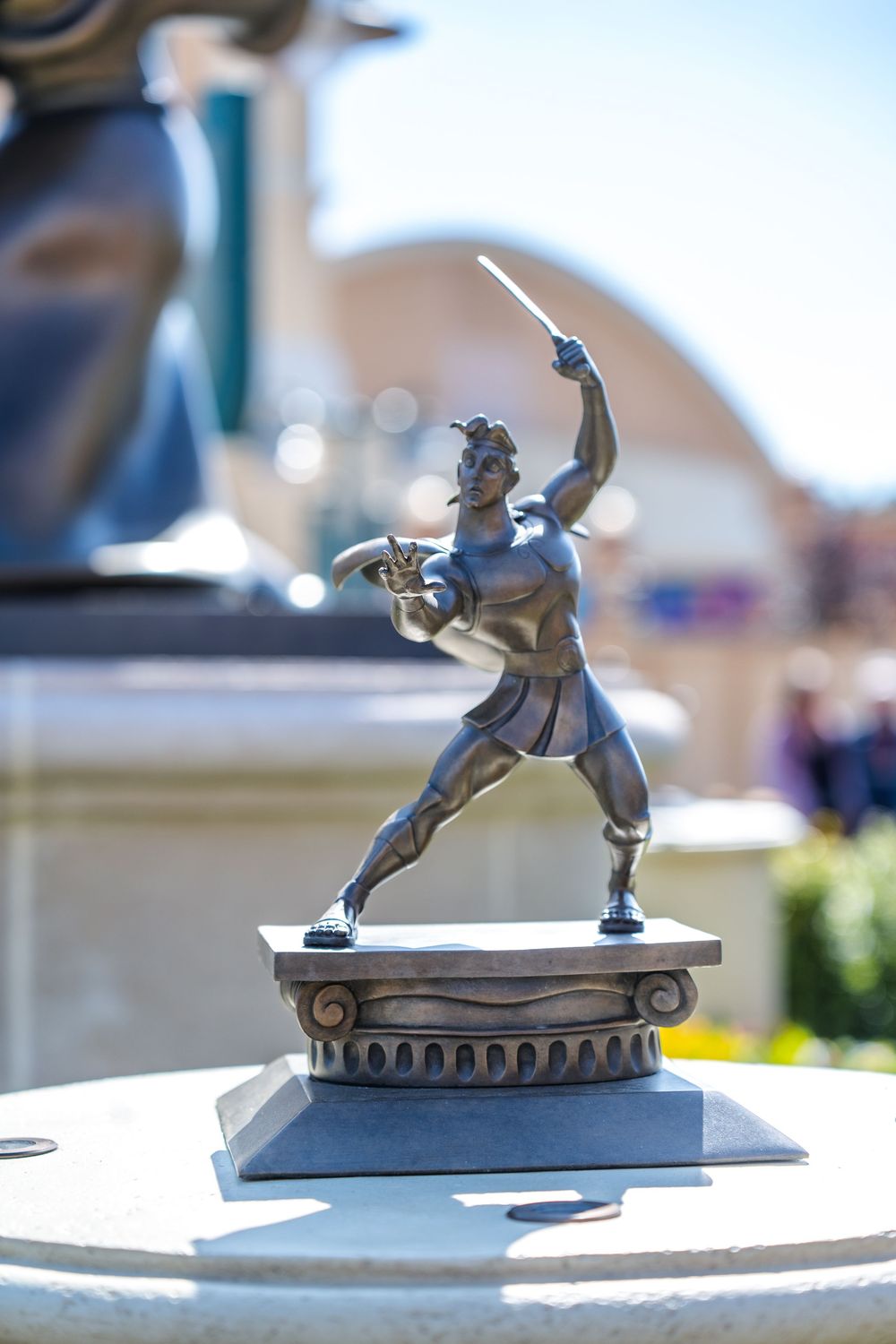 Small decorative bronze statue of an athlete in dynamic throwing pose atop ornate pedestal in outdoor setting.