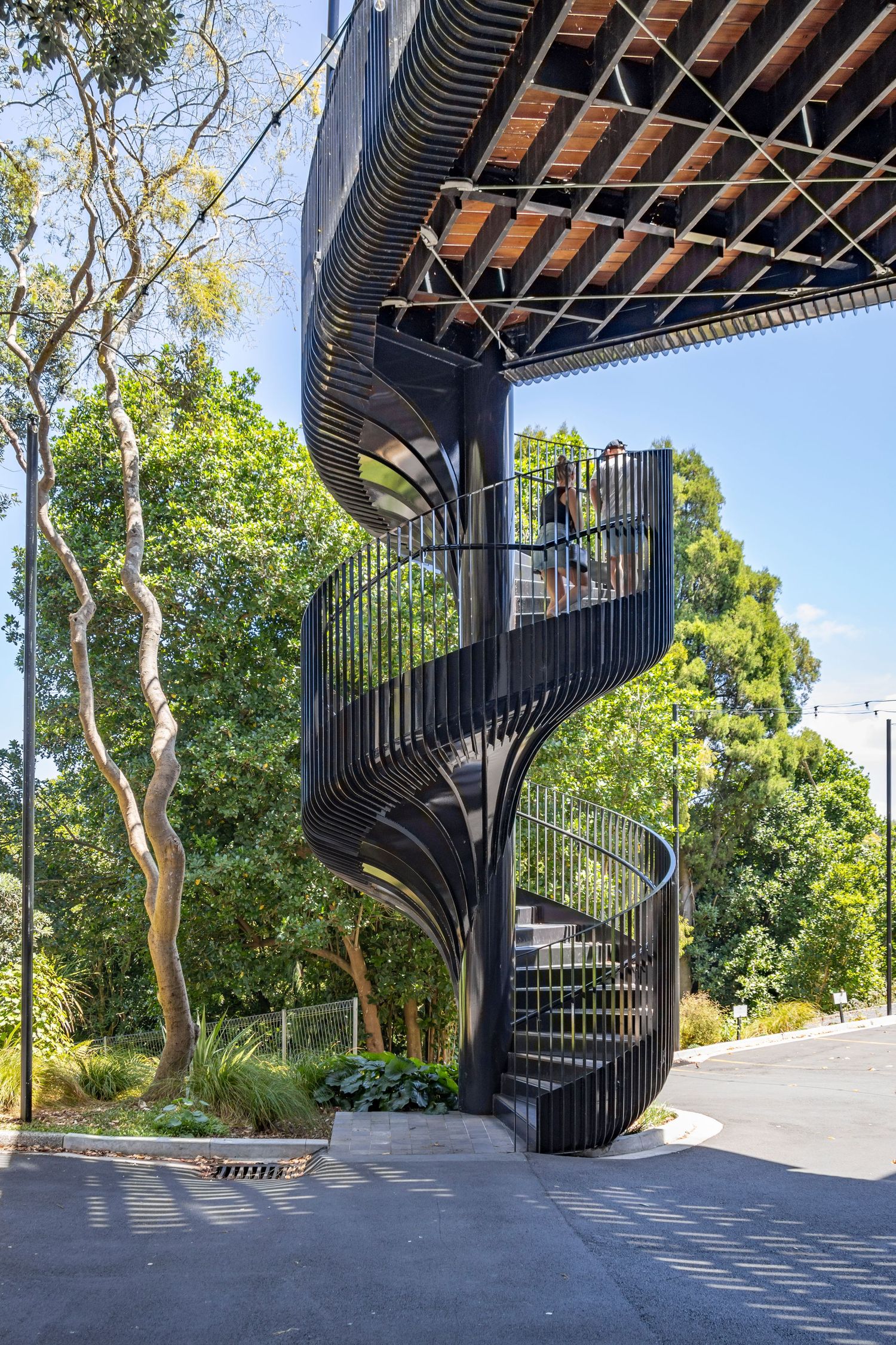 Black steel spiral staircase curls upward beneath a canopy, surrounded by lush green trees.