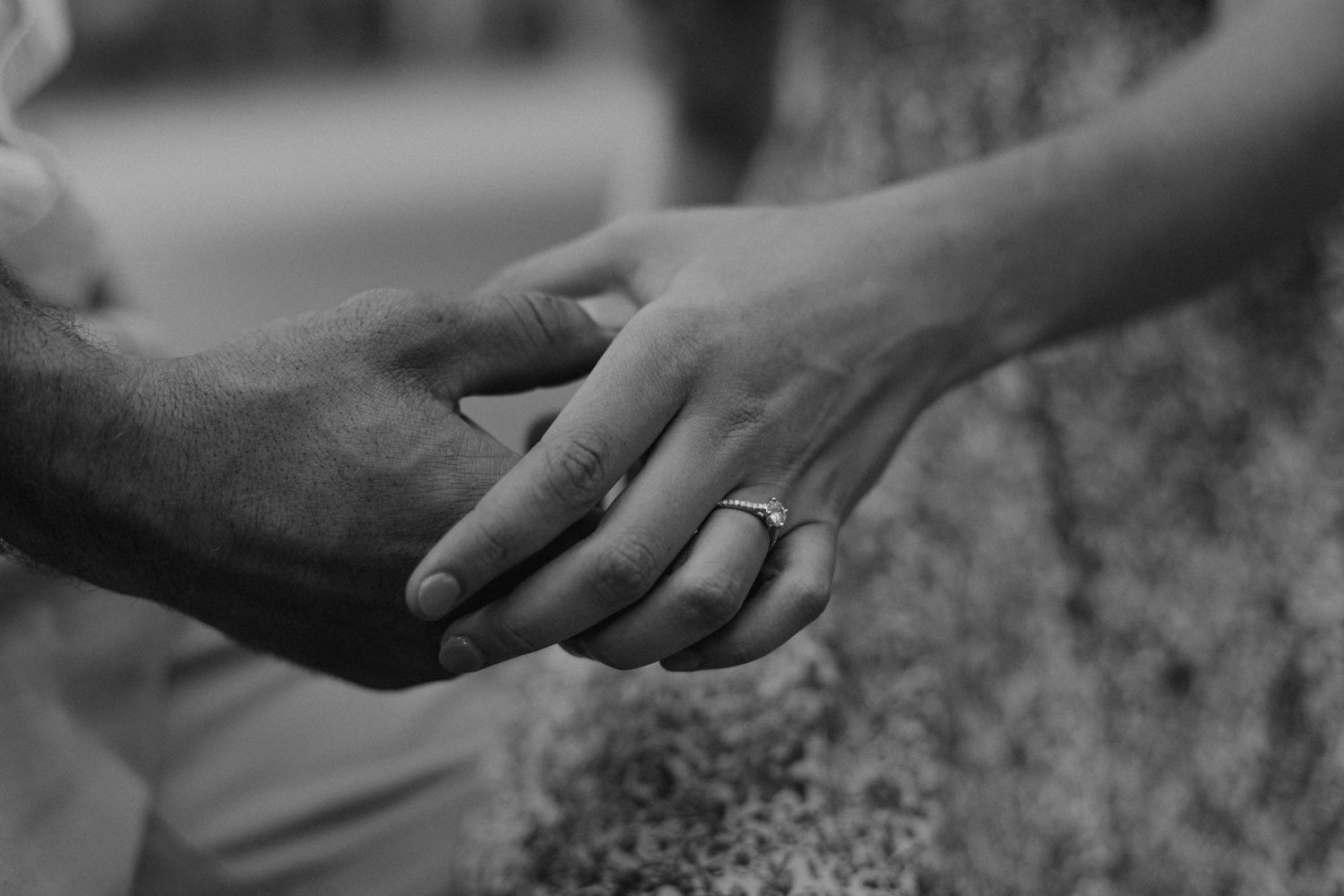 Close-up black and white photo of two hands with wedding rings touching against a textured surface.