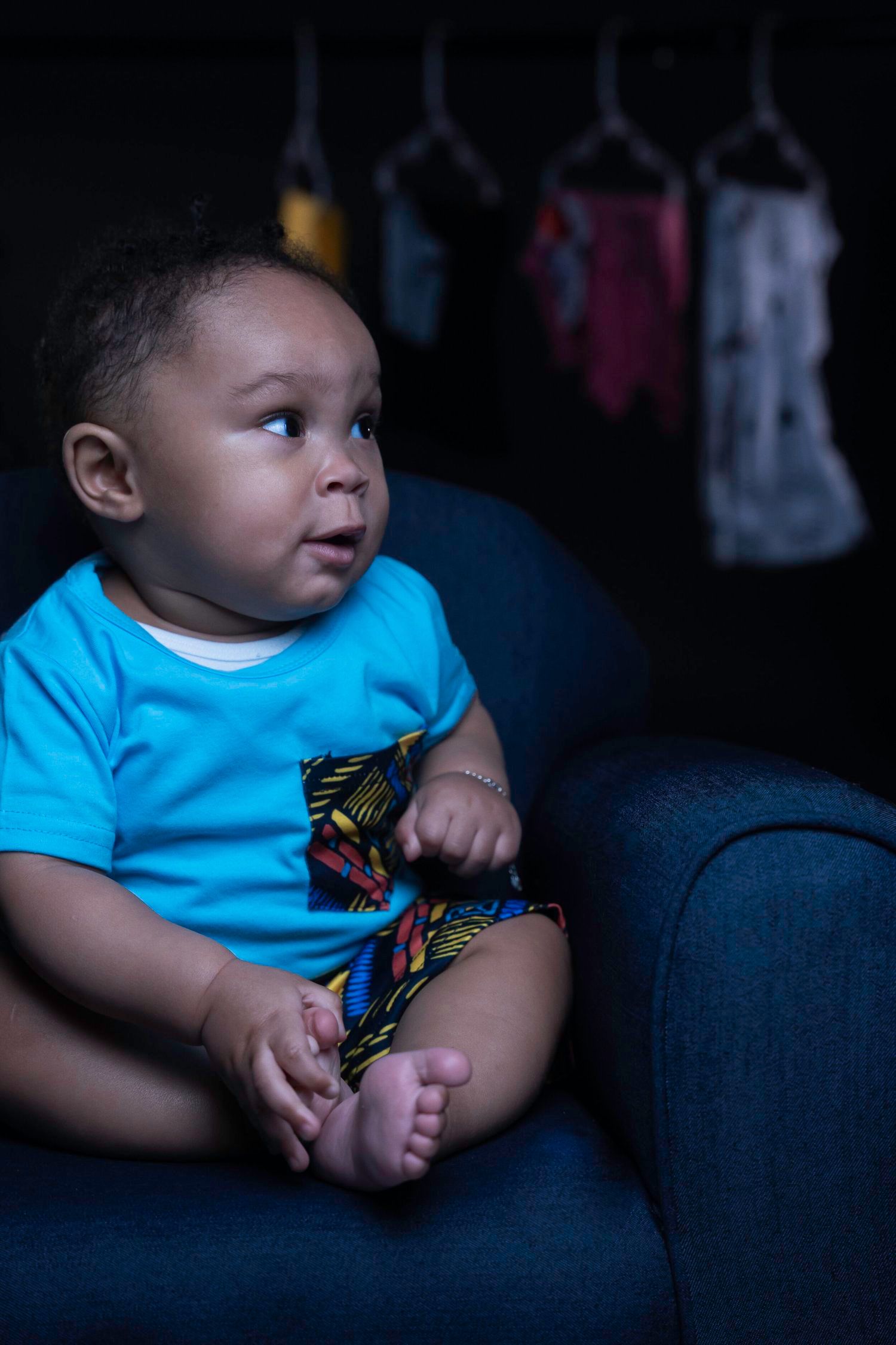 A young child in a bright blue shirt sits on a dark couch with hanging clothes visible in background.