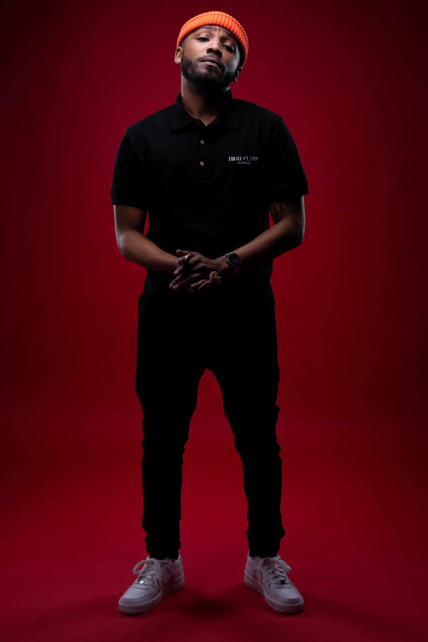 Fashion portrait against red backdrop showing person in black outfit and orange beanie.