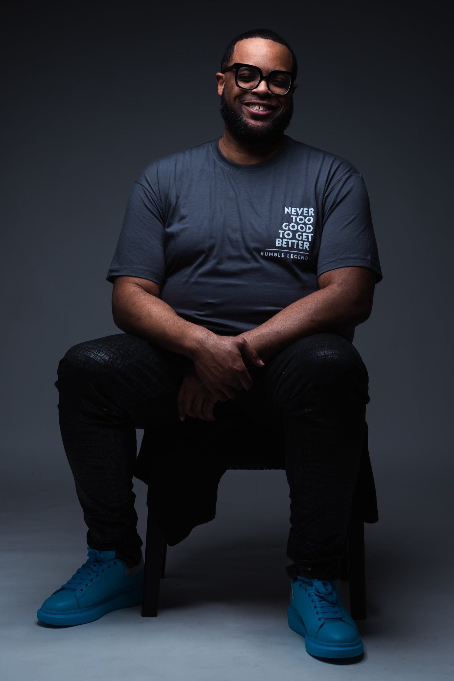 Studio portrait showing person seated wearing dark t-shirt and blue shoes.