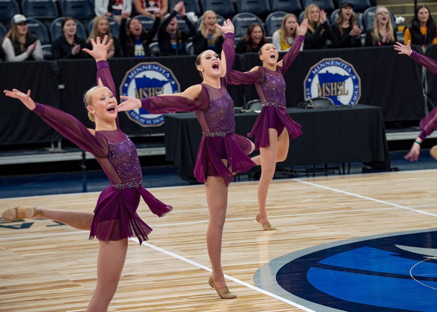 Dance team performs synchronized routine in purple costumes on wooden stage.