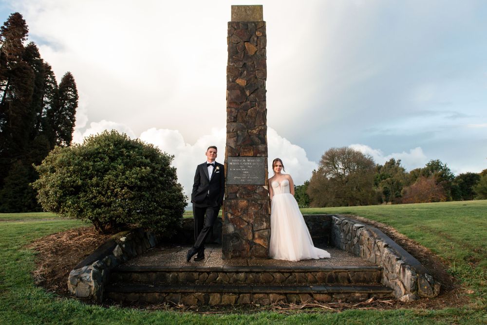 A wedding couple poses next to a tall stone pillar monument on landscaped grounds Cambridge New Zealand