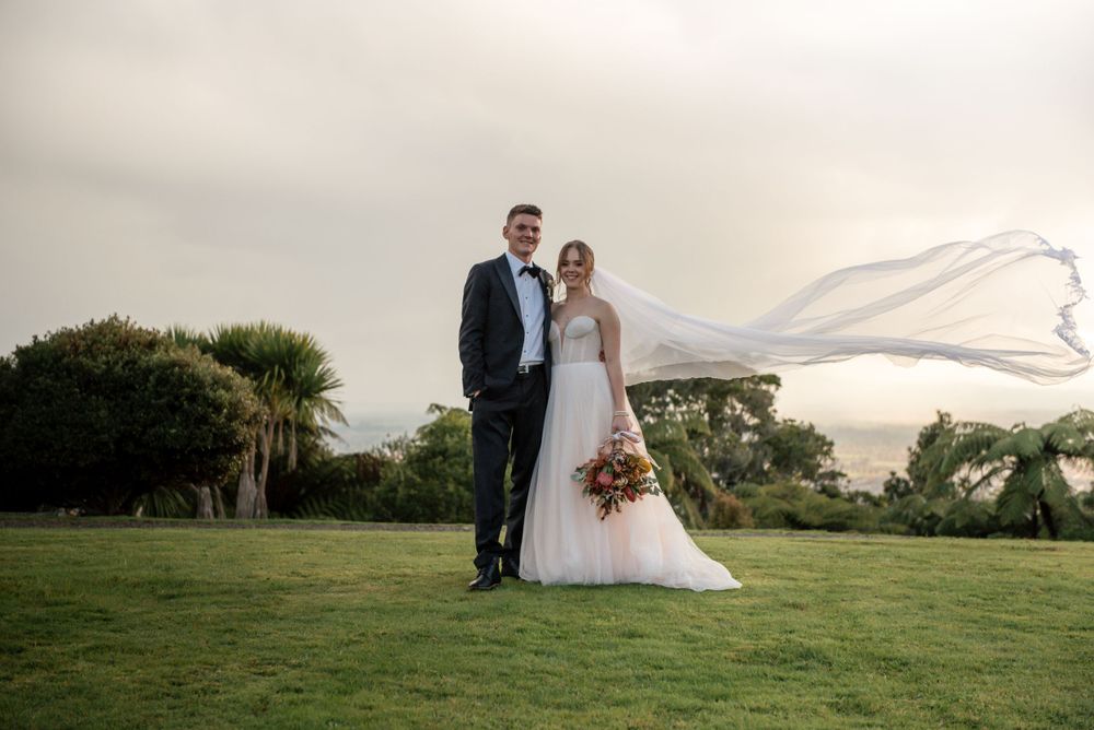 A wedding couple stands on a lush lawn as the bride's veil dramatically catches the wind Cambridge New Zealand
