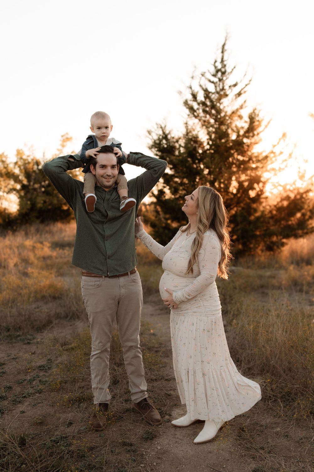 Family maternity photo shoot outdoors at golden hour with toddler sitting on shoulders and pregnant woman in white dress.