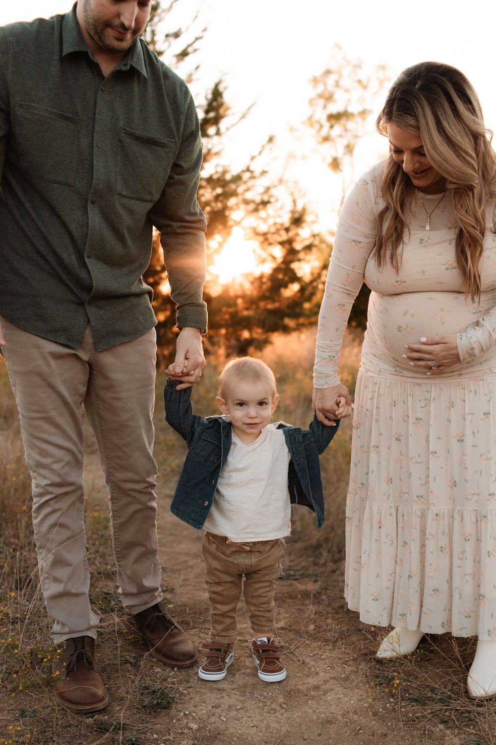 Sweet outdoor family photo with toddler holding parents' hands at sunset wearing casual clothing.