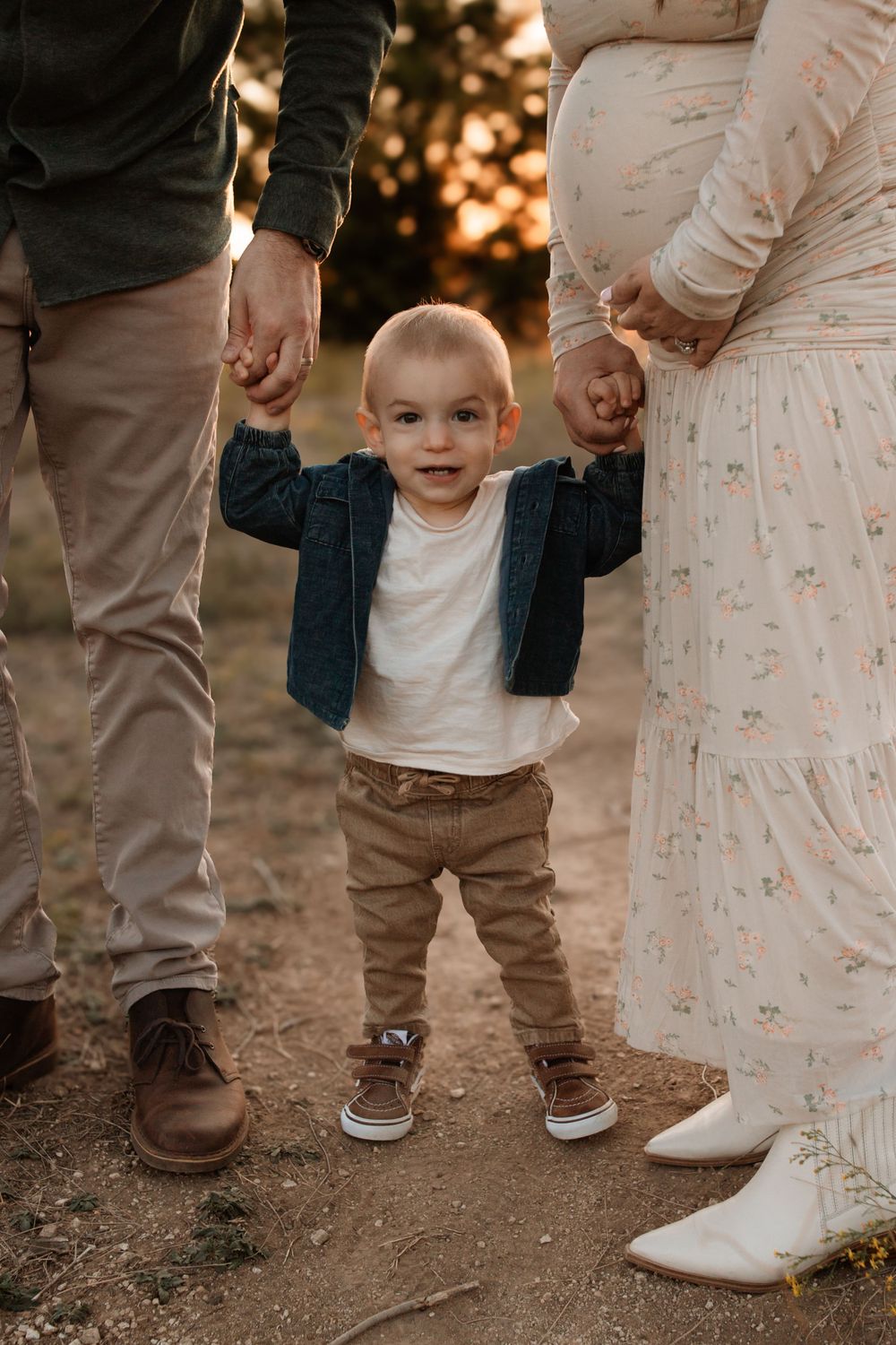 A toddler stands holding hands with parents during golden hour photoshoot.