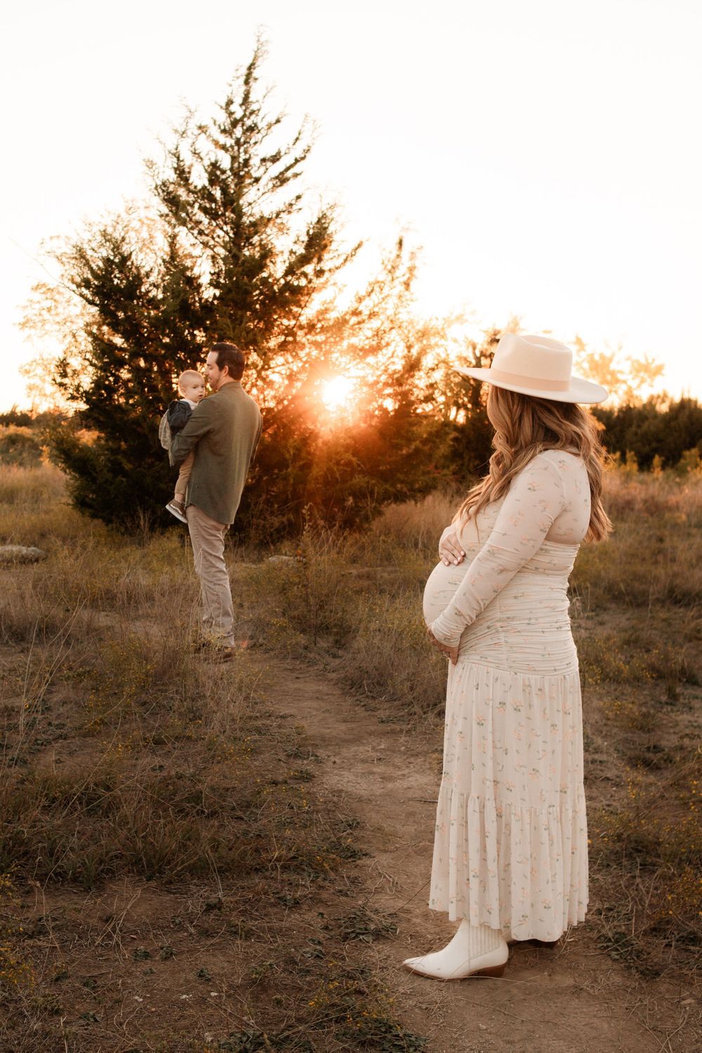 Maternity couple photo at sunset with woman in white dress and man standing near pine trees.