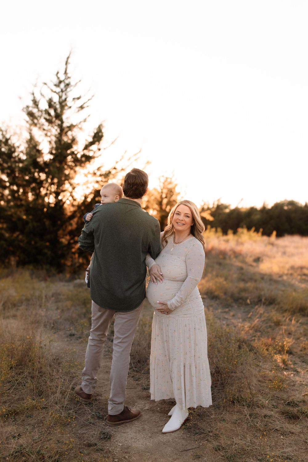 A couple stands together at sunset with the expectant mother in a flowing white dress against evergreen trees.