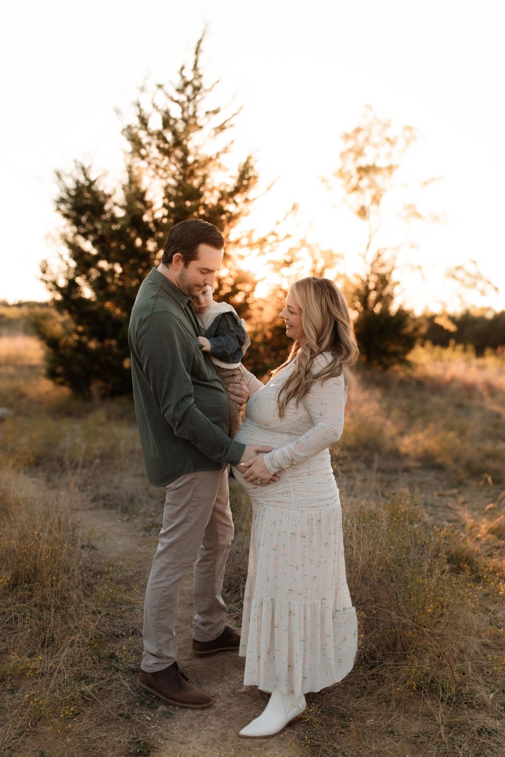 Expecting couple embraces with toddler during golden hour portrait session.
