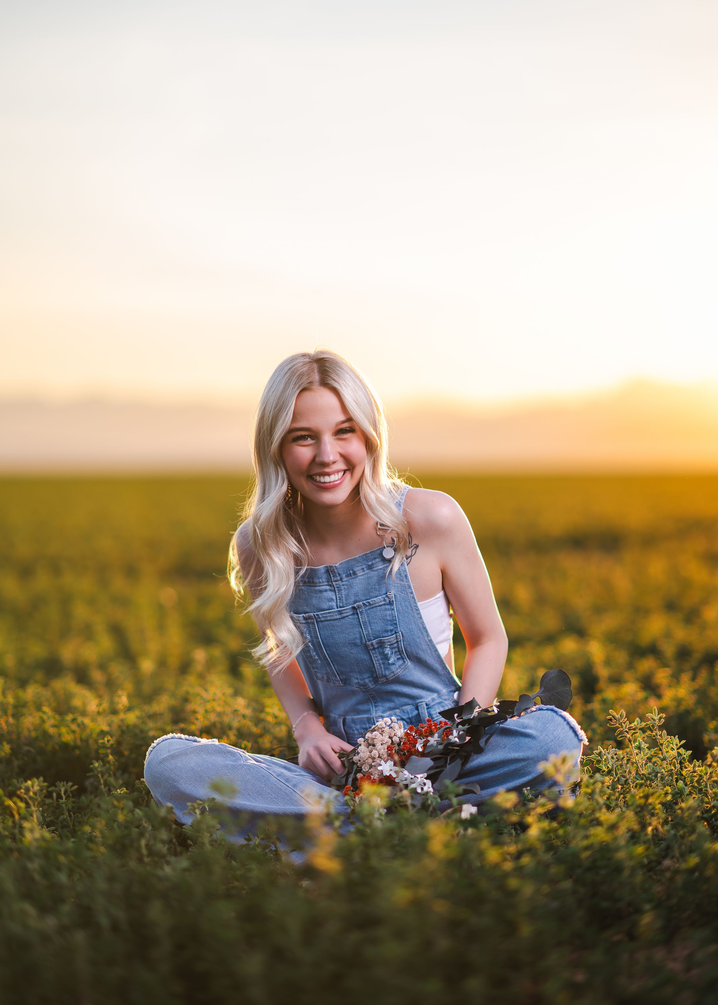 A person in denim overalls sits among blooming yellow flowers in a field during golden sunset hour. Senior portraits by Dusty Wooddell.