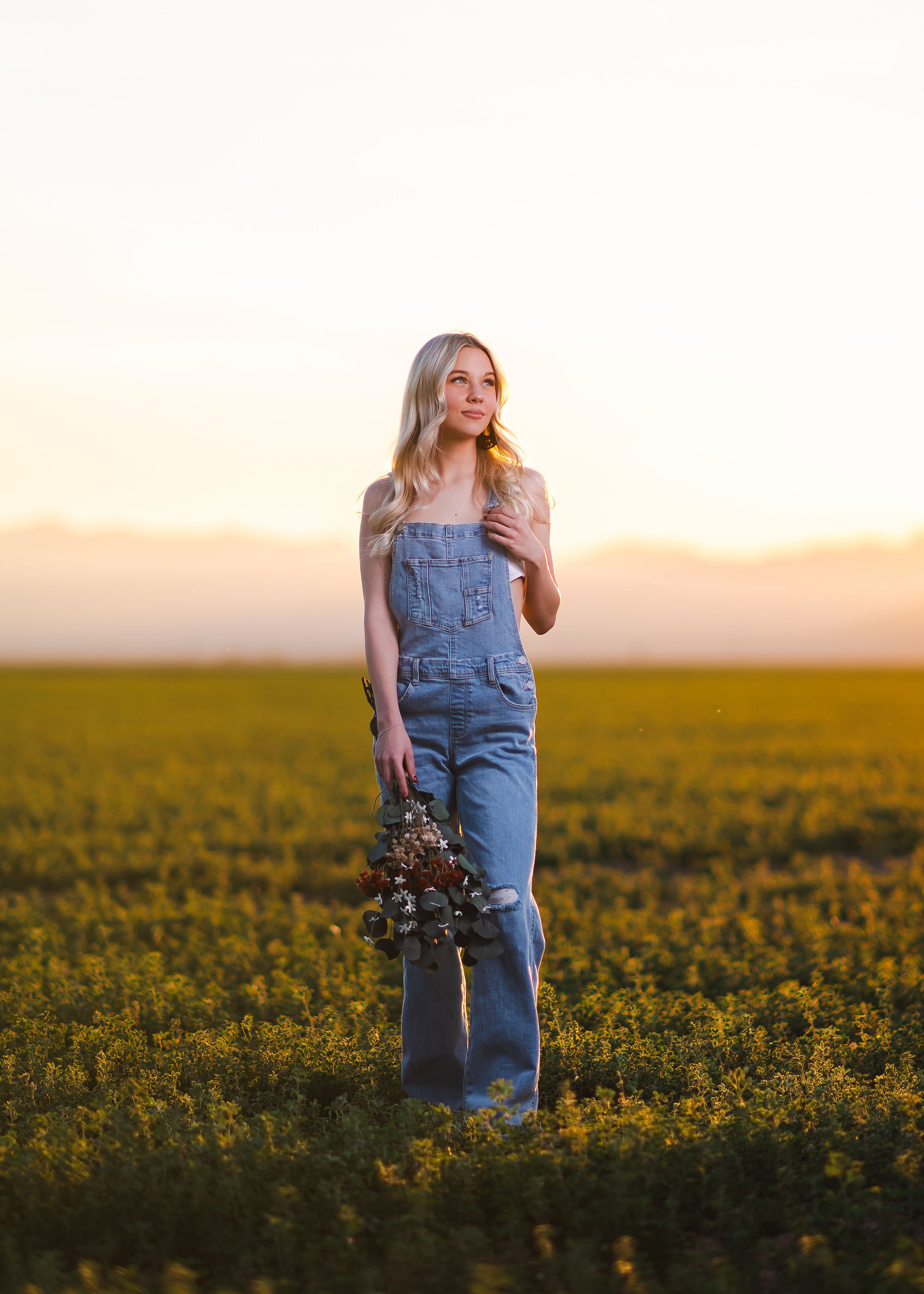 A person in denim overalls stands in a golden field of wildflowers at sunset holding a bouquet of dried flowers. Senior portraits by Dusty W