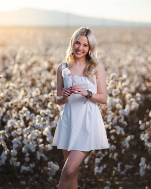 A woman in a white dress poses gracefully in a cotton field during golden hour lighting. Senior portraits by Dusty Wooddell.